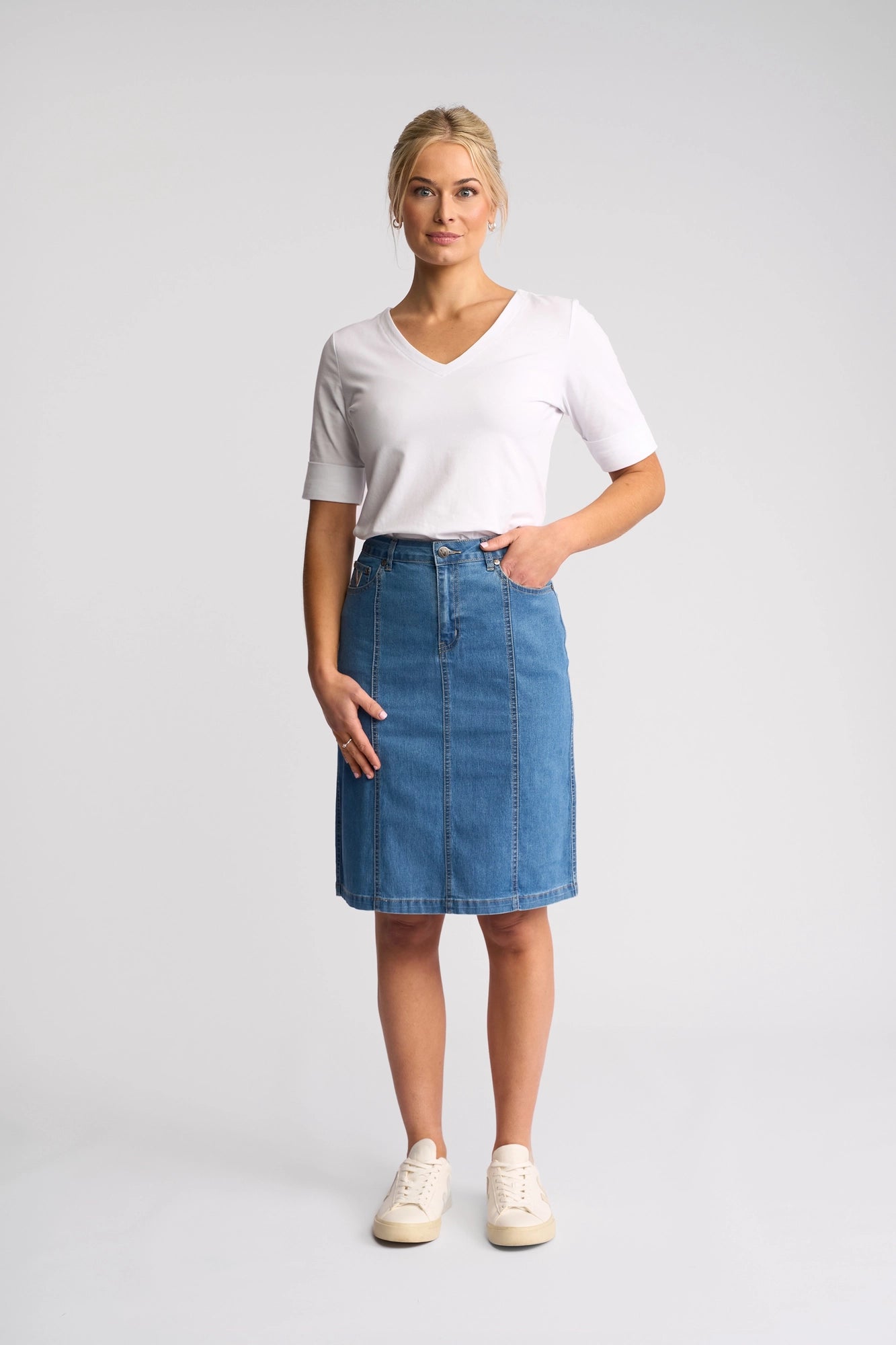A woman stands facing forward against a plain light background, wearing the Vassalli Poppy Skirt in Light Denim paired with a white short-sleeve V-neck shirt and white sneakers, with one hand in her skirt pocket.