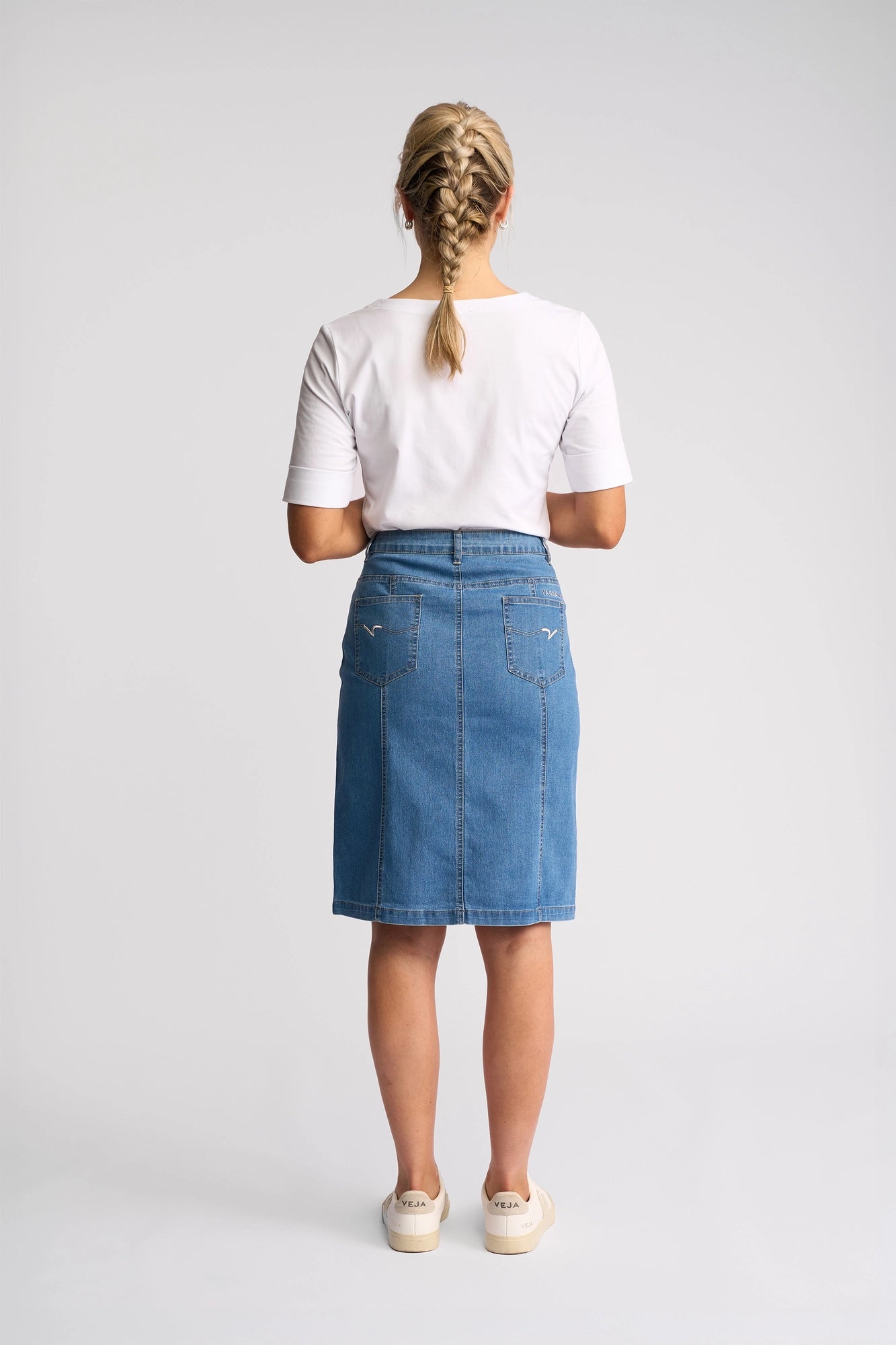 A woman with blonde braided hair stands facing away against a plain light background, wearing a white short-sleeve shirt, white sneakers, and the Vassalli Poppy Skirt in Light Denim.