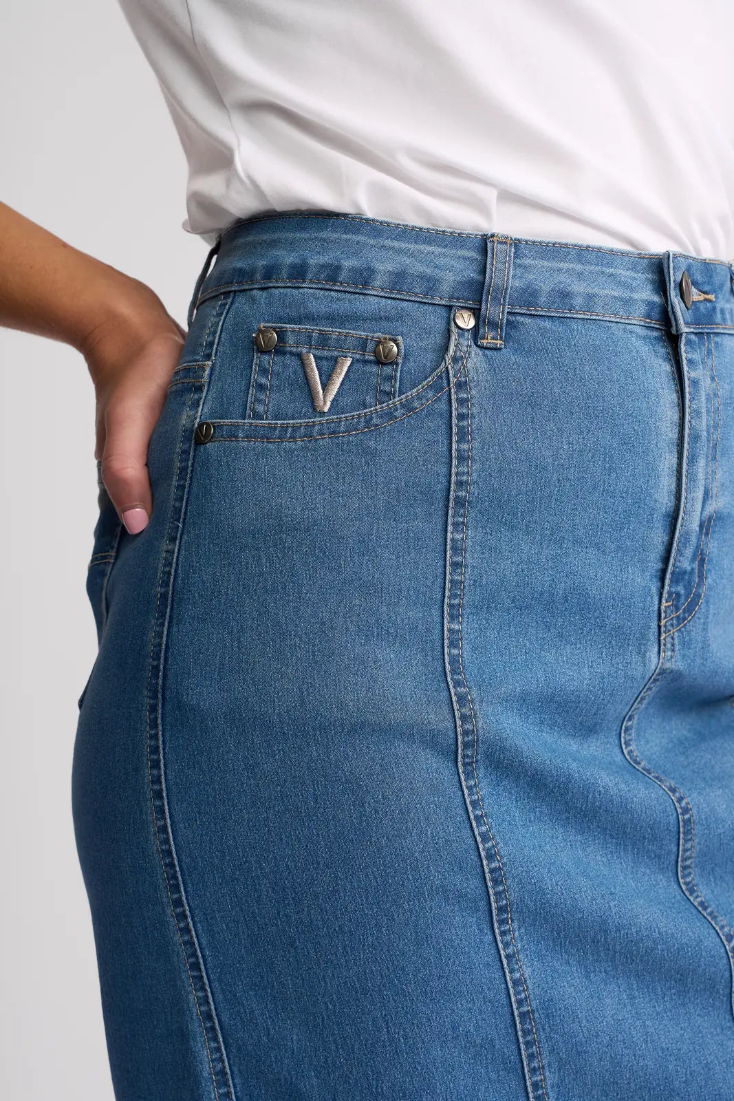A close-up of a person wearing the Vassalli Poppy Skirt in light denim paired with a white shirt, with one hand on their hip. The summer skirt features visible stitching and a small pocket adorned with a metal V detail.