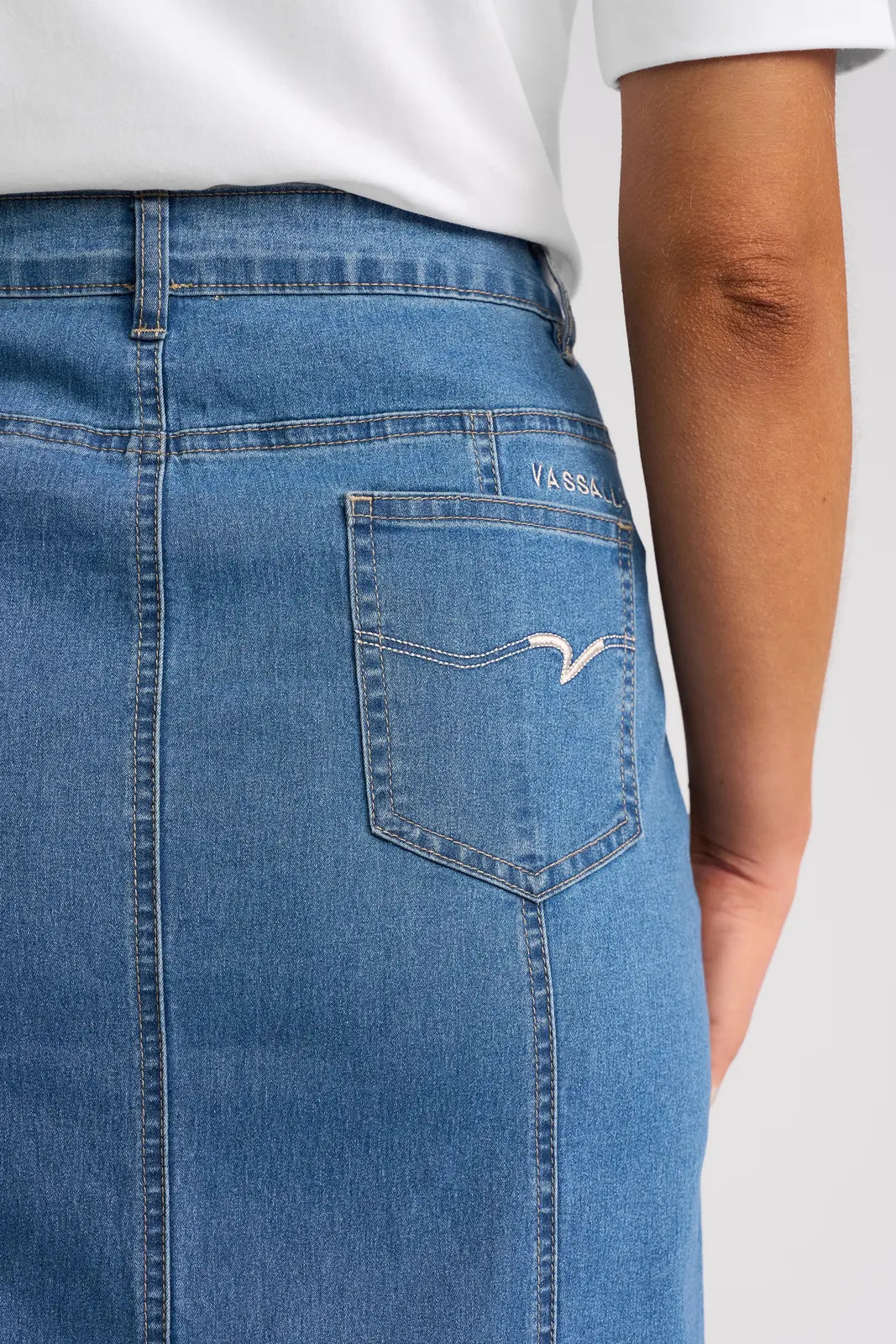 Close-up of a person wearing the Vassalli Poppy Skirt in light denim, featuring white stitching on the back pocket and a small Vassalli label. The person is also wearing a white top.
