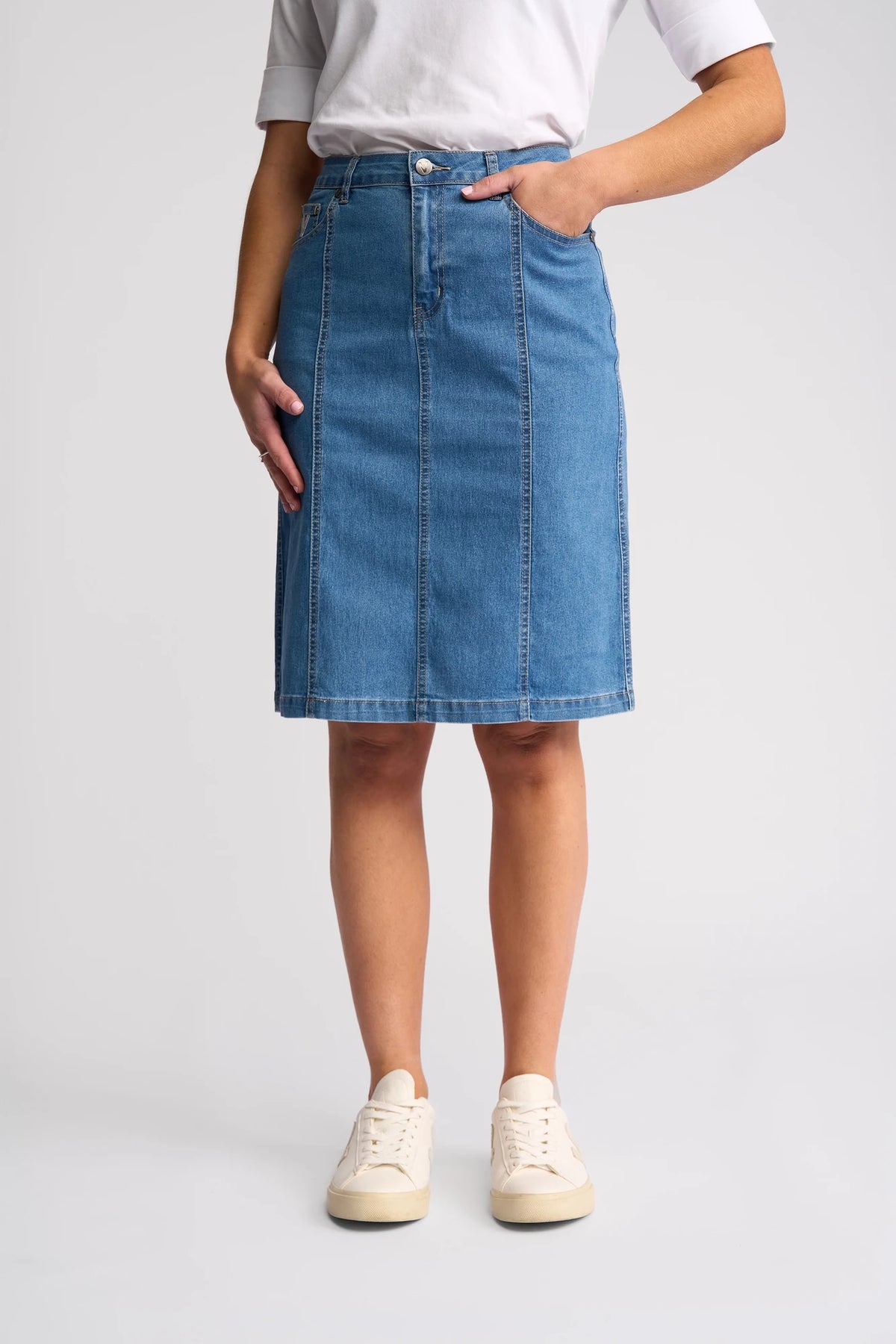 A person wearing a white t-shirt, the Vassalli Poppy Skirt in Light Denim—a knee-length pencil skirt—and white sneakers, stands with one hand in a pocket against a plain background.