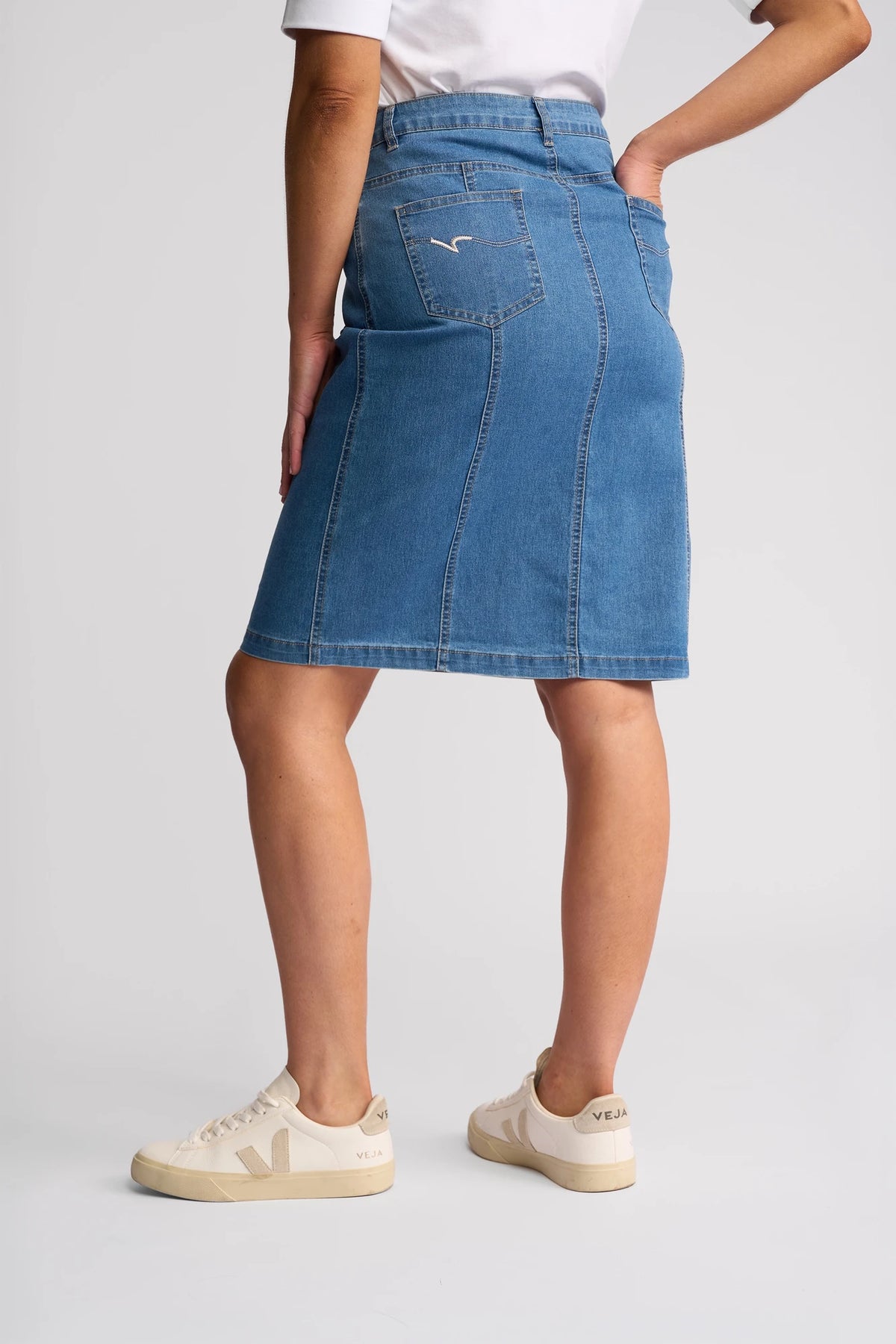 A person wearing the Vassalli Poppy Skirt in Light Denim, paired with white Veja sneakers and a white shirt, stands against a plain light background facing away from the camera.