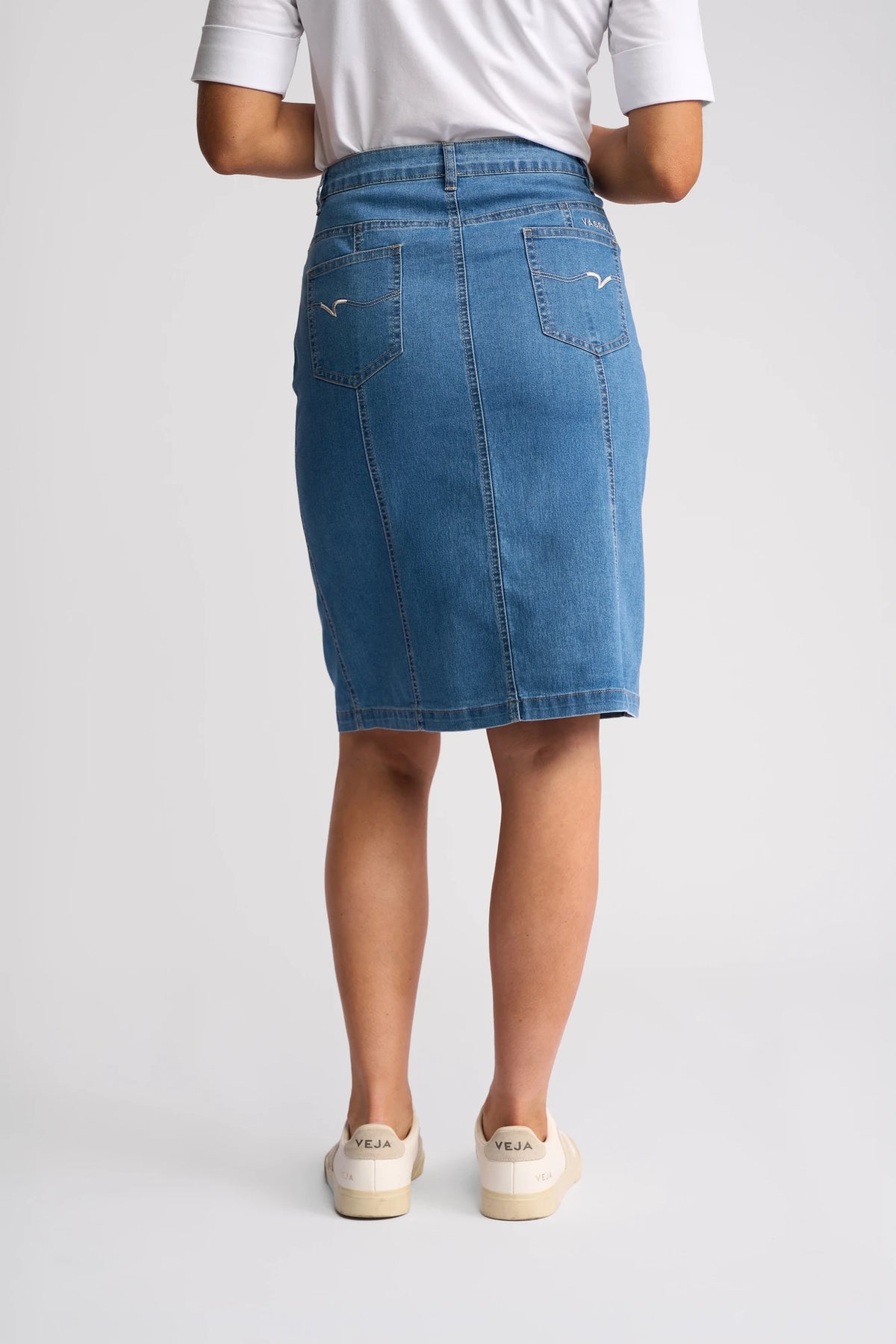 A person stands facing away, wearing a white shirt and the Vassalli Poppy Skirt in Light Denim, which features back pockets and a pencil silhouette. They have paired the look with beige Veja sneakers against a plain, light-colored background.