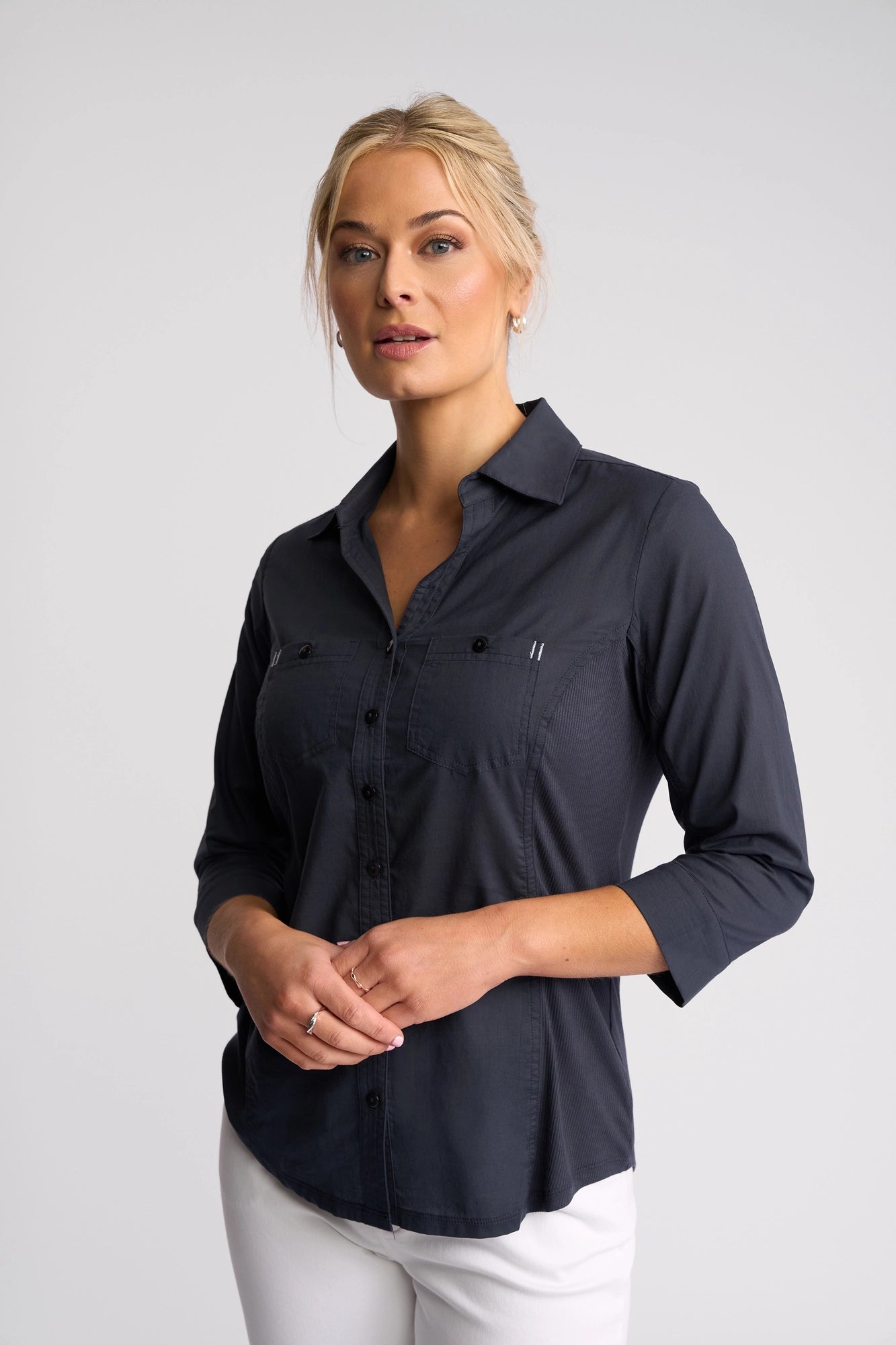 A woman with blond hair in a bun wears the Vassalli Isla Shirt in Ink, a dark, lightweight cotton summer shirt with 3/4 length sleeves, paired with white pants and standing against a plain light background.