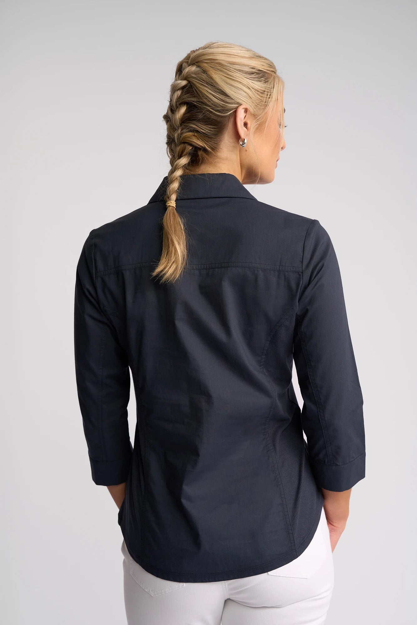 A woman with blonde hair in a braid is shown from the back, wearing the Vassalli Isla Shirt in Ink—a dark long-sleeve top featuring a ribbed fabric panel—paired with white pants, standing against a plain light background.