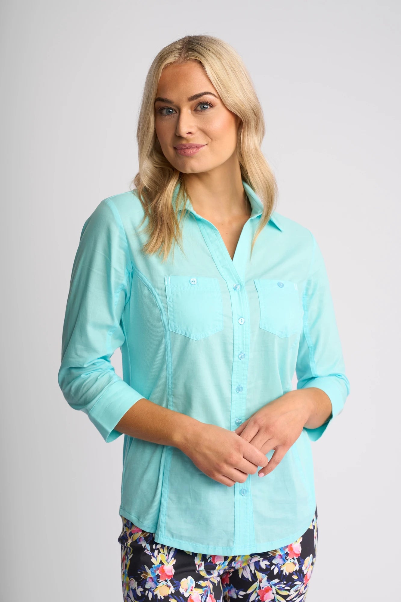 A woman with blonde hair wears the Vassalli Isla Shirt in Marine—a light blue, lightweight cotton summer shirt featuring front pockets—paired with dark floral pants, standing against a plain light gray background.
