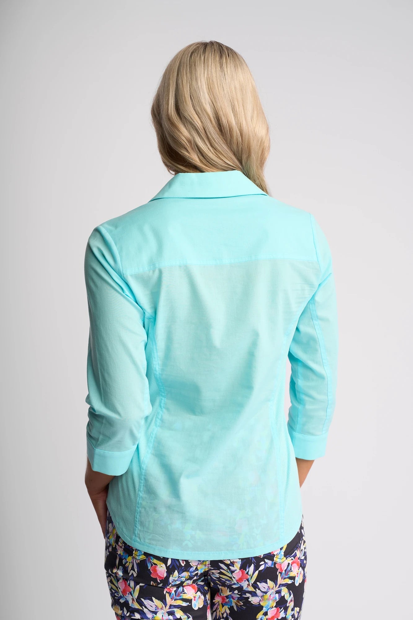 A person with long blonde hair is shown from the back, wearing the Vassalli Isla Shirt in Marine and dark floral pants. The background is plain and light-colored.