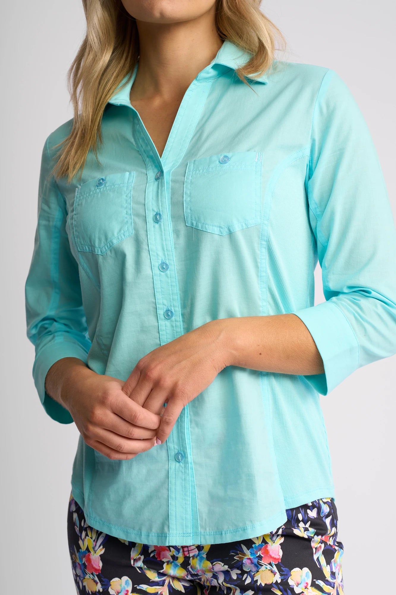 A woman wears the Vassalli Isla Shirt in Marine—a light blue, lightweight cotton summer shirt with front pockets—paired with navy pants adorned with a vibrant floral pattern. Only her torso and arms are visible, hands gently clasped, set against a plain light-colored background.