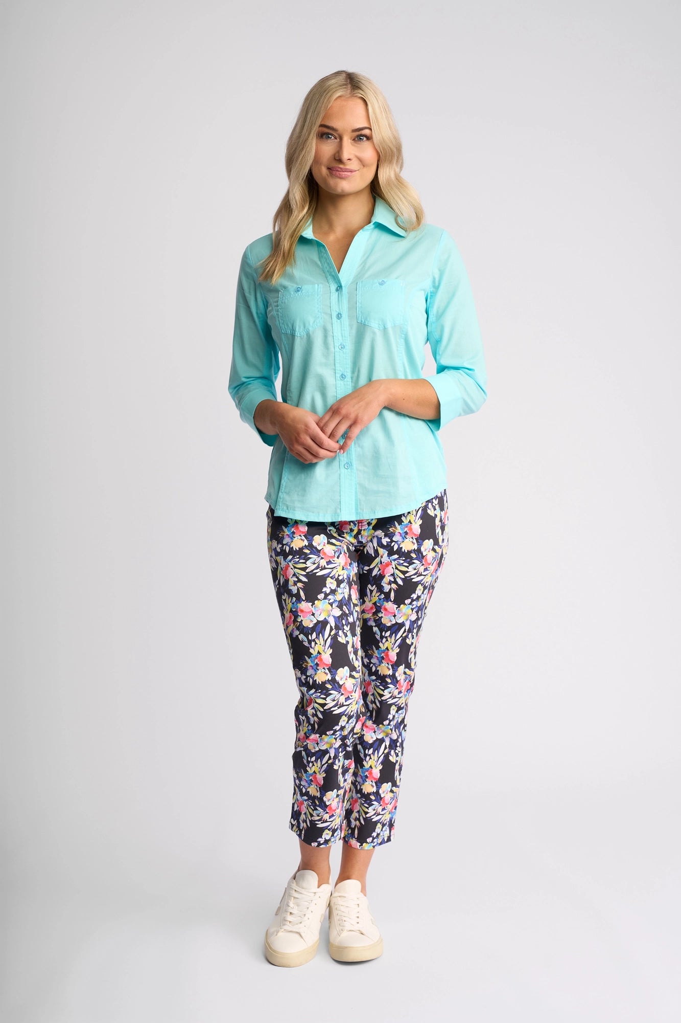 A woman with long blonde hair wears a Vassalli Isla Shirt in Marine, floral print pants, and white sneakers, standing and smiling against a plain light gray background.