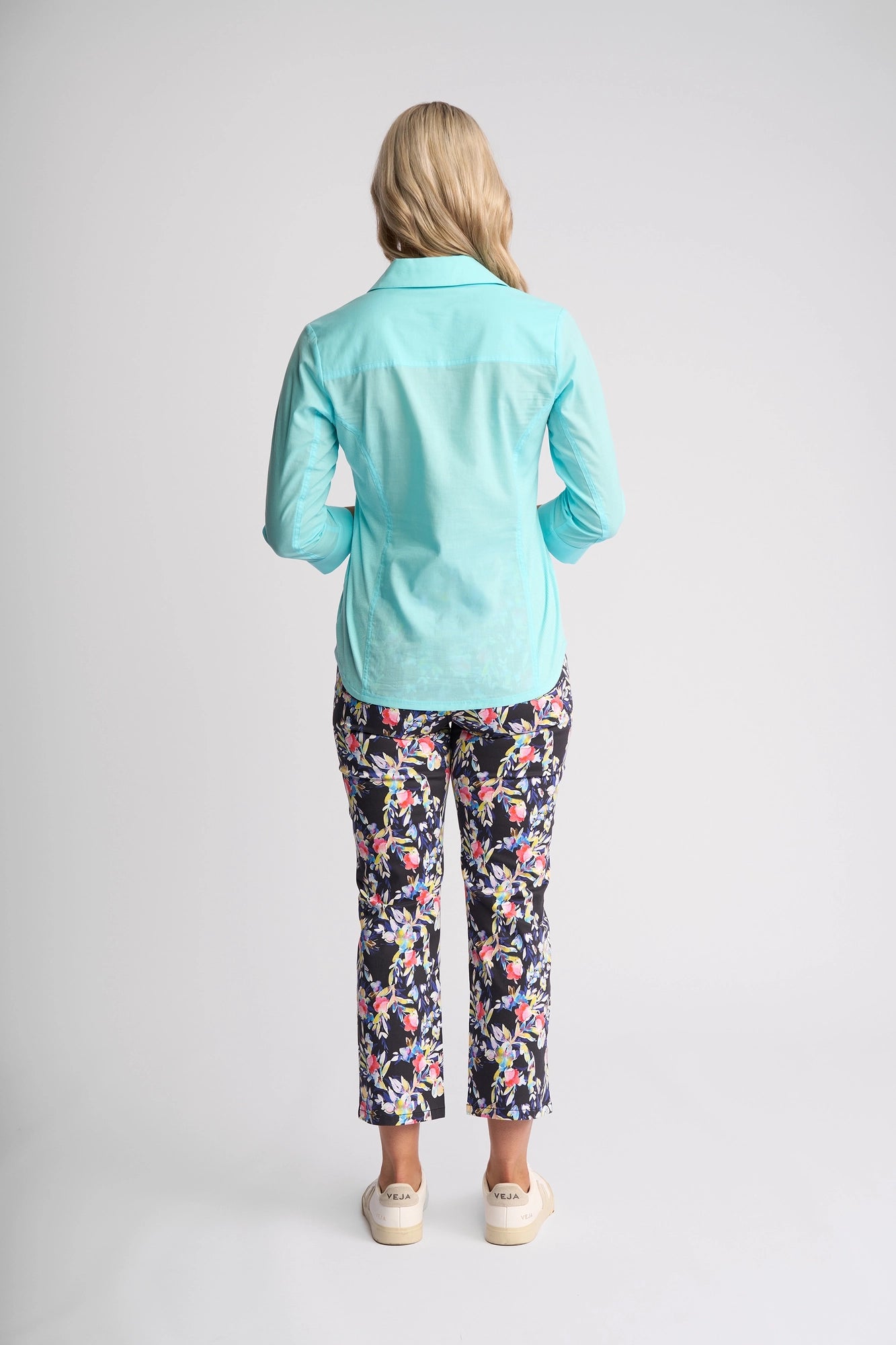 A woman stands facing away, wearing the Vassalli Isla Shirt in Marine—a light blue, lightweight cotton summer shirt—paired with floral patterned pants and white shoes, set against a plain white background.
