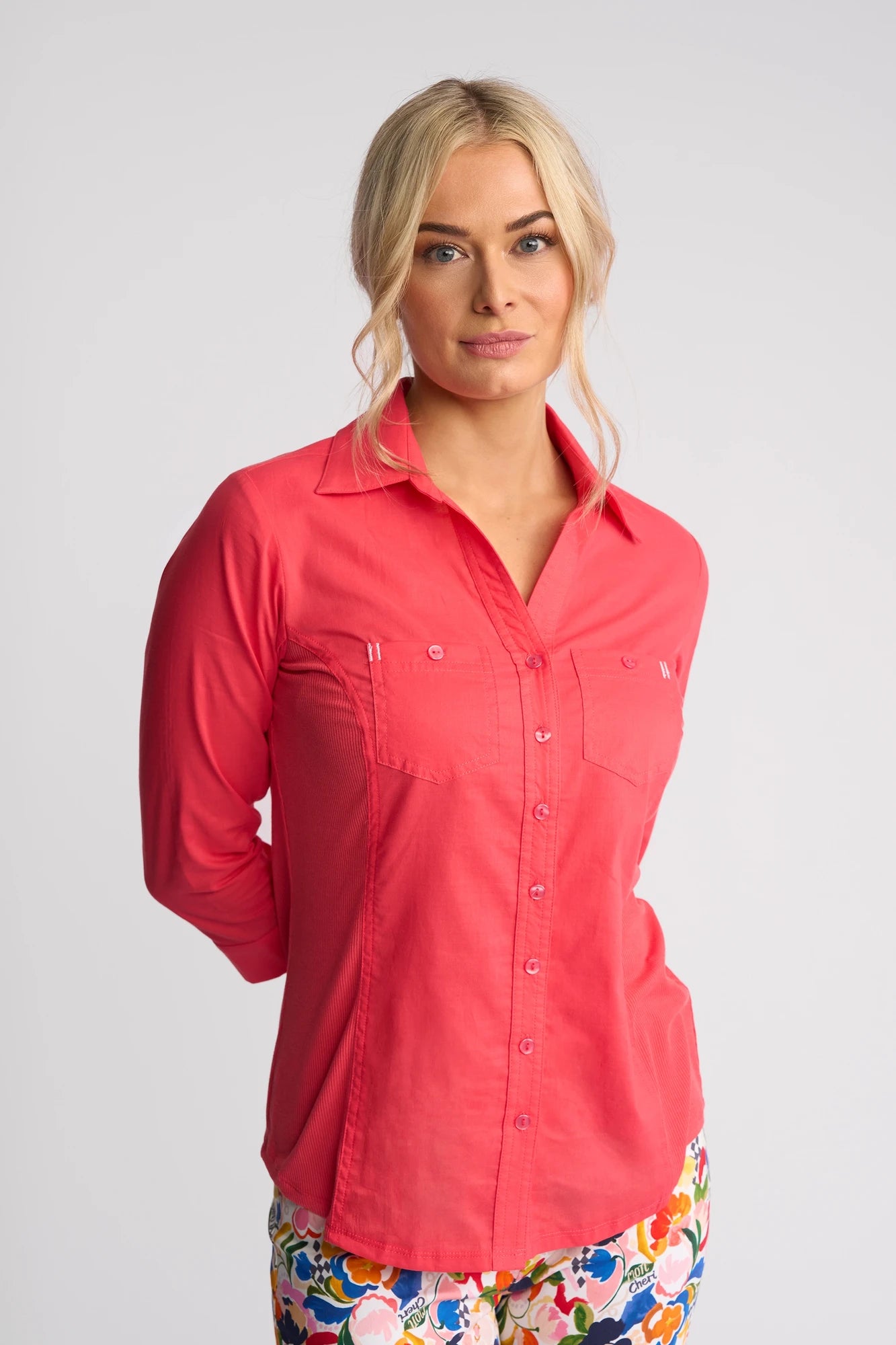 A blonde woman wearing a vibrant Vassalli Isla Shirt - Raspberry and colorful floral pants stands against a plain, light gray background with her hands behind her back.