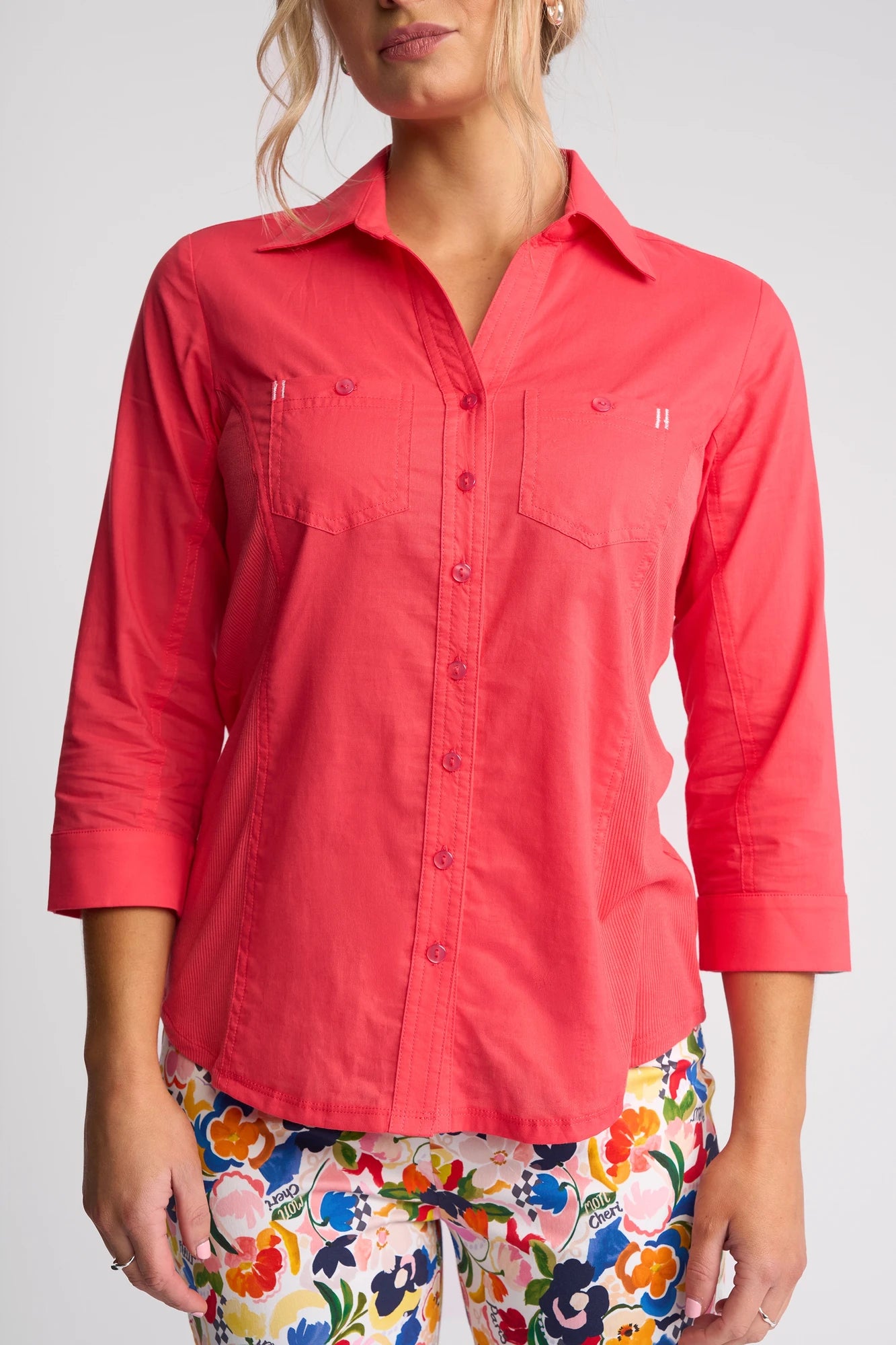 A woman wearing the Vassalli Isla Shirt in Raspberry with three-quarter sleeves and vibrant floral pants stands against a plain light background, her face partially out of frame.