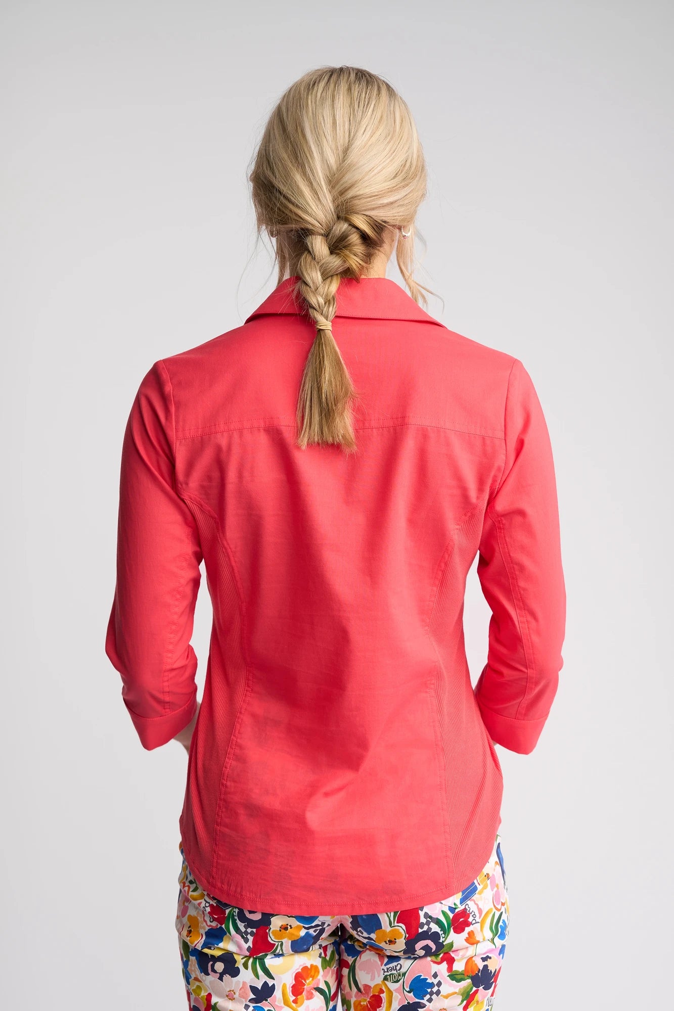 A woman with blond hair in a braid is seen from behind, wearing the Vassalli Isla Shirt in Raspberry and colorful floral pants against a plain light background.