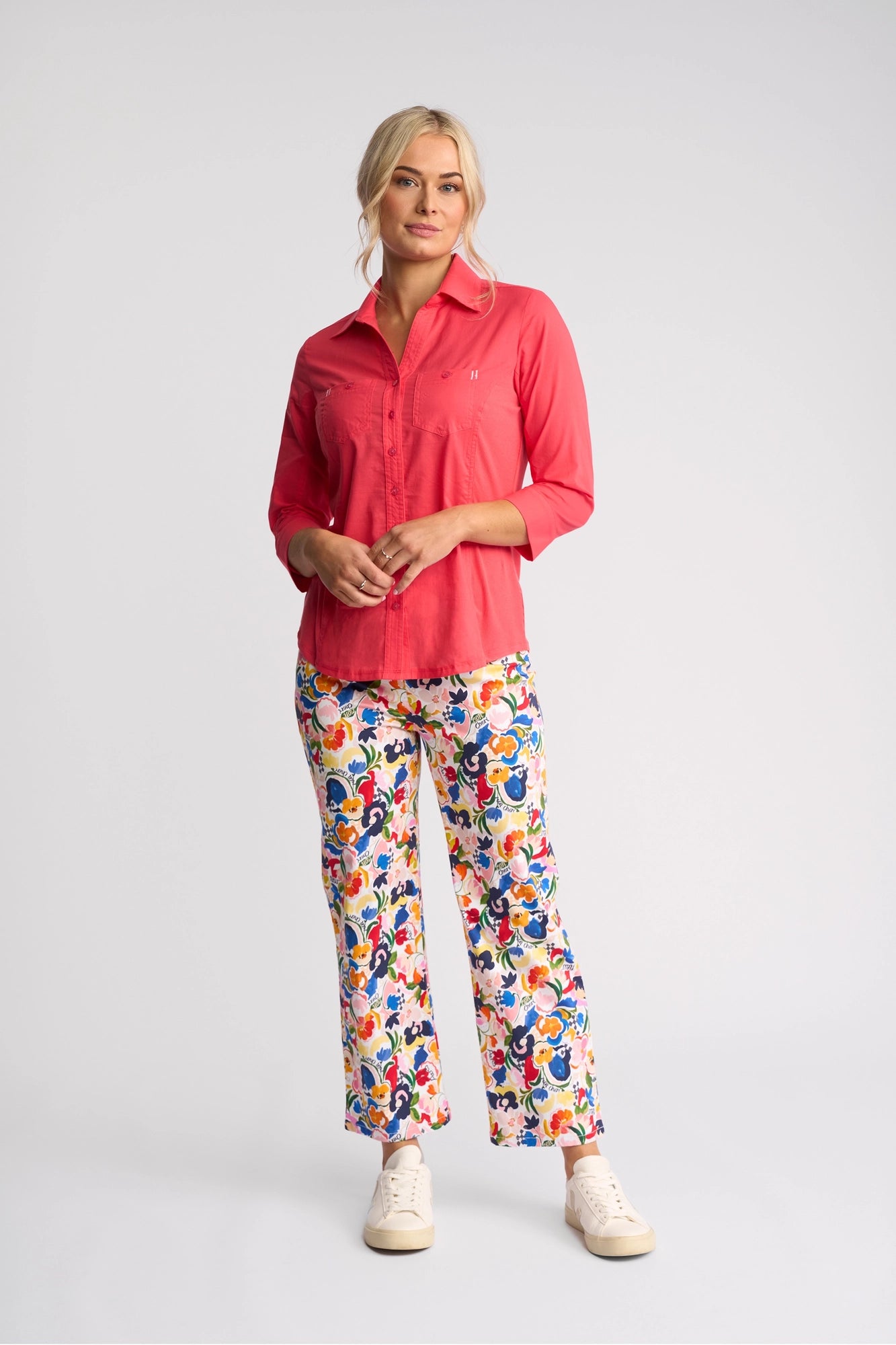 A woman stands against a plain background, wearing the Vassalli Isla Shirt in Raspberry paired with colorful floral print pants and white sneakers. Her blonde hair is tied back, and she has a relaxed, confident expression, perfectly showcasing this lightweight shirt from Vassalli—an ideal choice for summer days.