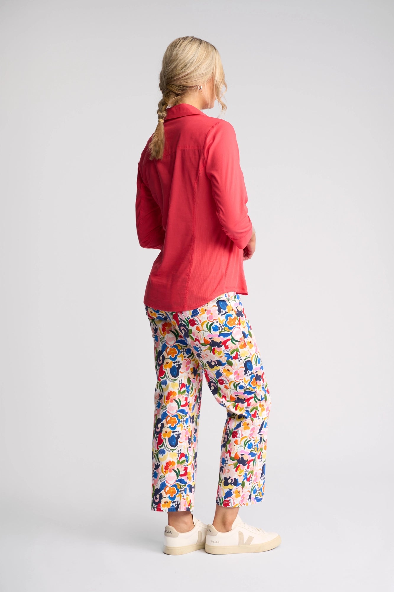 A person with blonde hair in a braid wears a long-sleeve Vassalli Isla Shirt in Raspberry, colorful floral pants, and beige sneakers, standing against a plain light background and facing away from the camera.