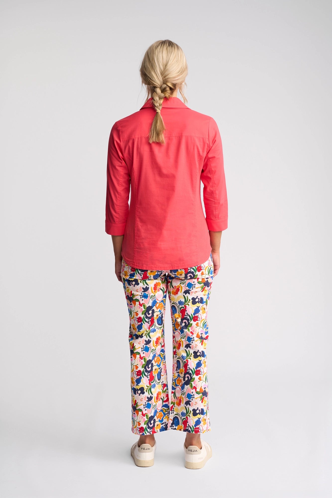 A person with blonde hair in a braid, wearing the vibrant Vassalli Isla Shirt in Raspberry, stands facing away from the camera against a light gray background.