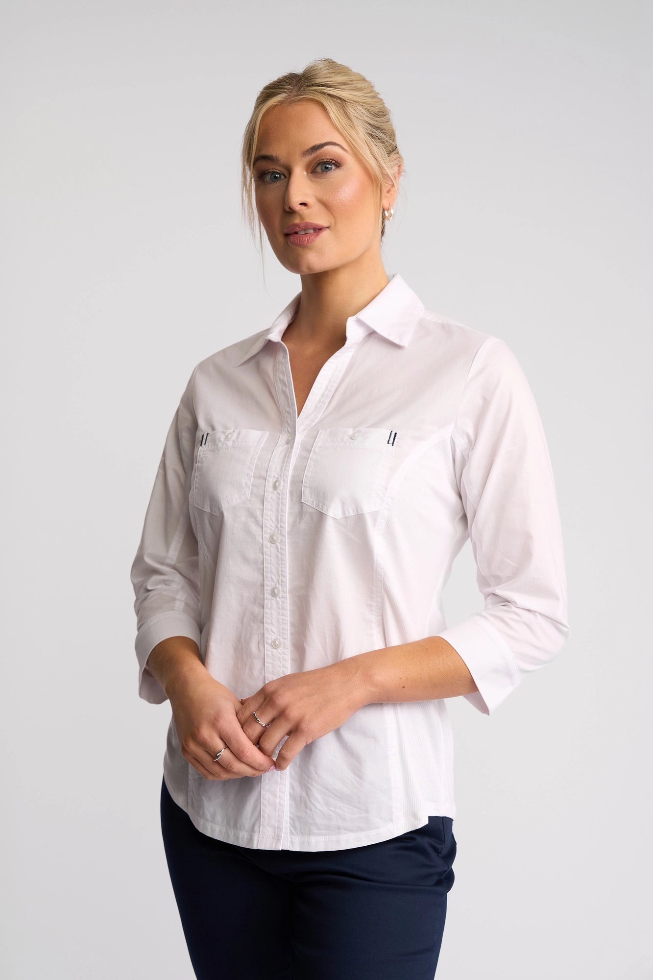 A woman with blonde hair styled in a neat updo wears the Vassalli Isla Shirt - White, featuring 3/4 sleeves, paired with dark pants and poses against a plain light background.
