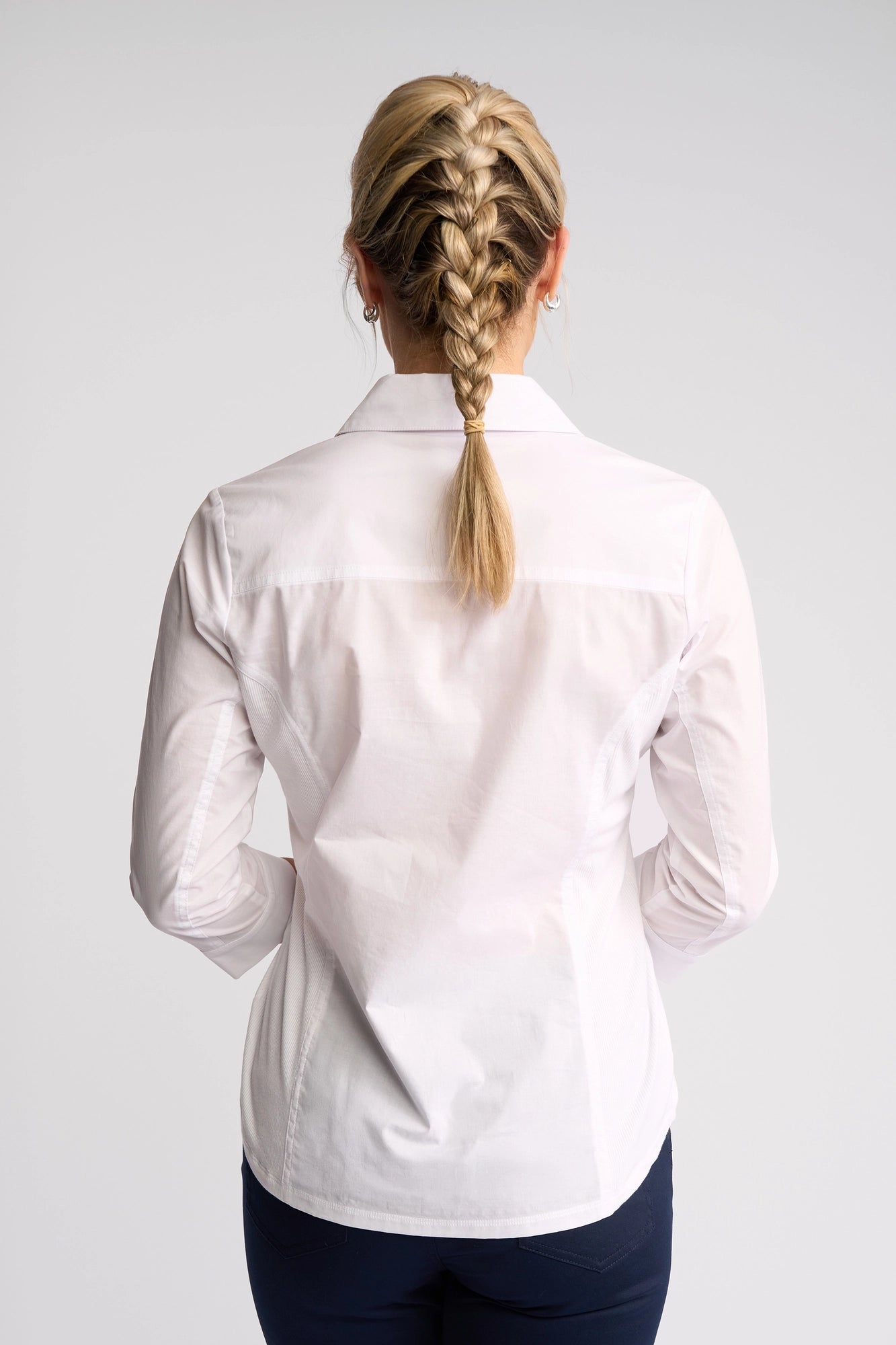 A person with blonde hair styled in a French braid stands with their back to the camera, dressed in the Vassalli Isla Shirt - White and dark pants against a simple, light background. This lightweight cotton shirt from Vassalli delivers an effortlessly chic summer style.