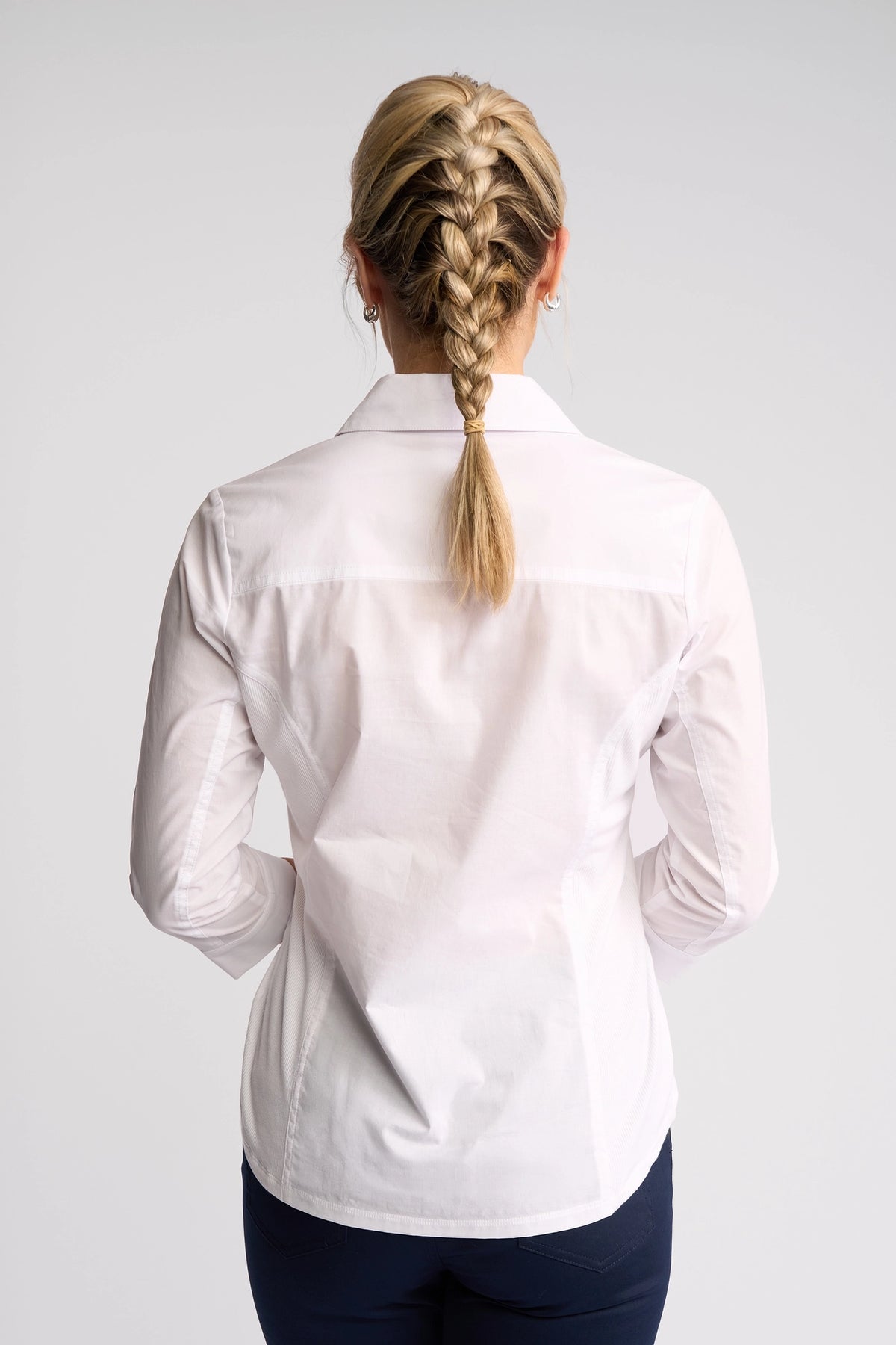 A person with blonde hair styled in a French braid stands with their back to the camera, dressed in the Vassalli Isla Shirt - White and dark pants against a simple, light background. This lightweight cotton shirt from Vassalli delivers an effortlessly chic summer style.