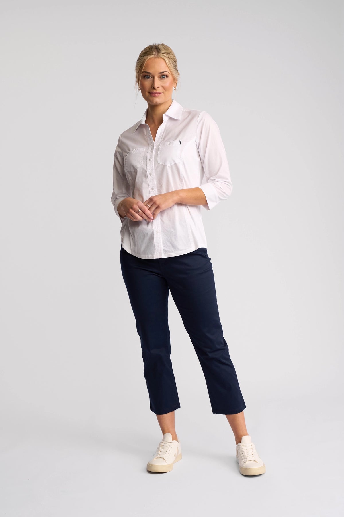 A woman with blonde hair styled back stands against a plain background, looking forward with a neutral expression. She is wearing the Vassalli Isla Shirt in white, paired with dark navy cropped pants and white sneakers.