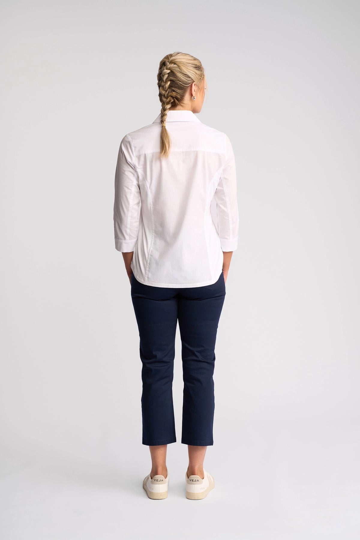 A person with blonde hair in braids stands facing away, wearing the Vassalli Isla Shirt in white, paired with navy pants and white shoes against a plain light background.