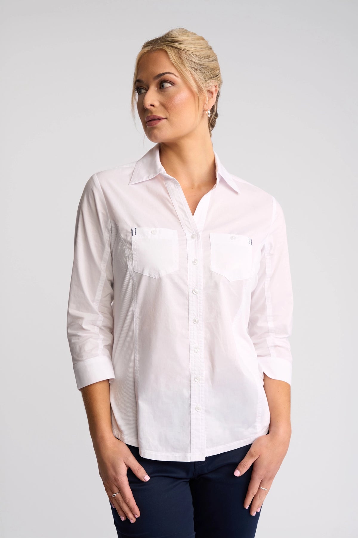 A woman stands against a plain background, wearing the Vassalli Isla Shirt in white with rolled-up sleeves and navy pants. Her blonde hair is styled up, and she looks to the side with a slight smile.