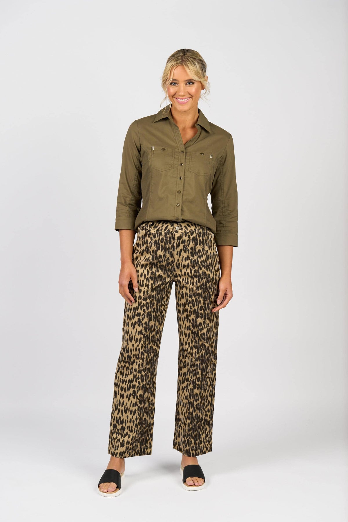 A woman smiles against a plain white background, wearing the Vassalli Isla Shirt in olive with three-quarter sleeves, paired with leopard print wide-leg pants and black sandals.