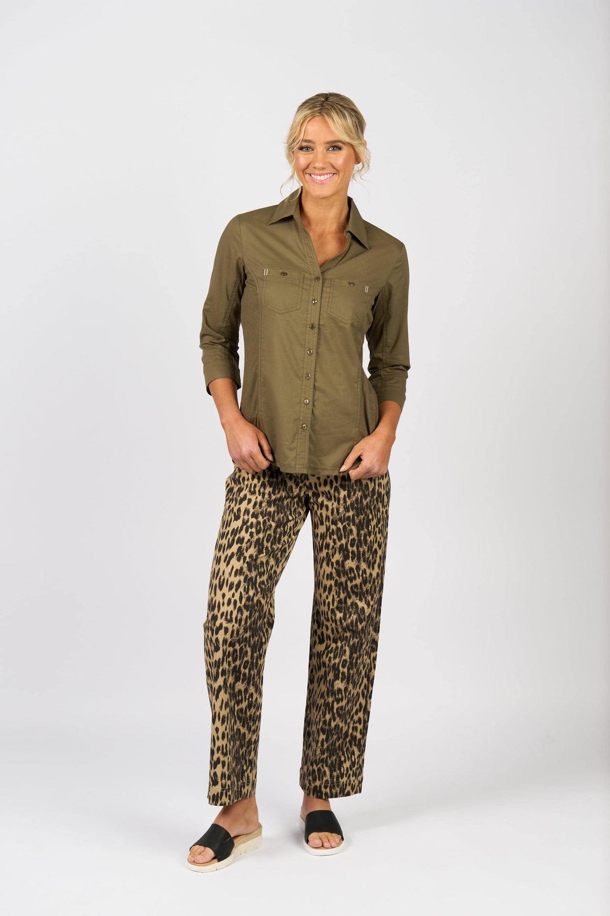 A woman smiles in front of a white background, wearing the Vassalli Isla Shirt in olive with wide-leg leopard print pants and black slide sandals.