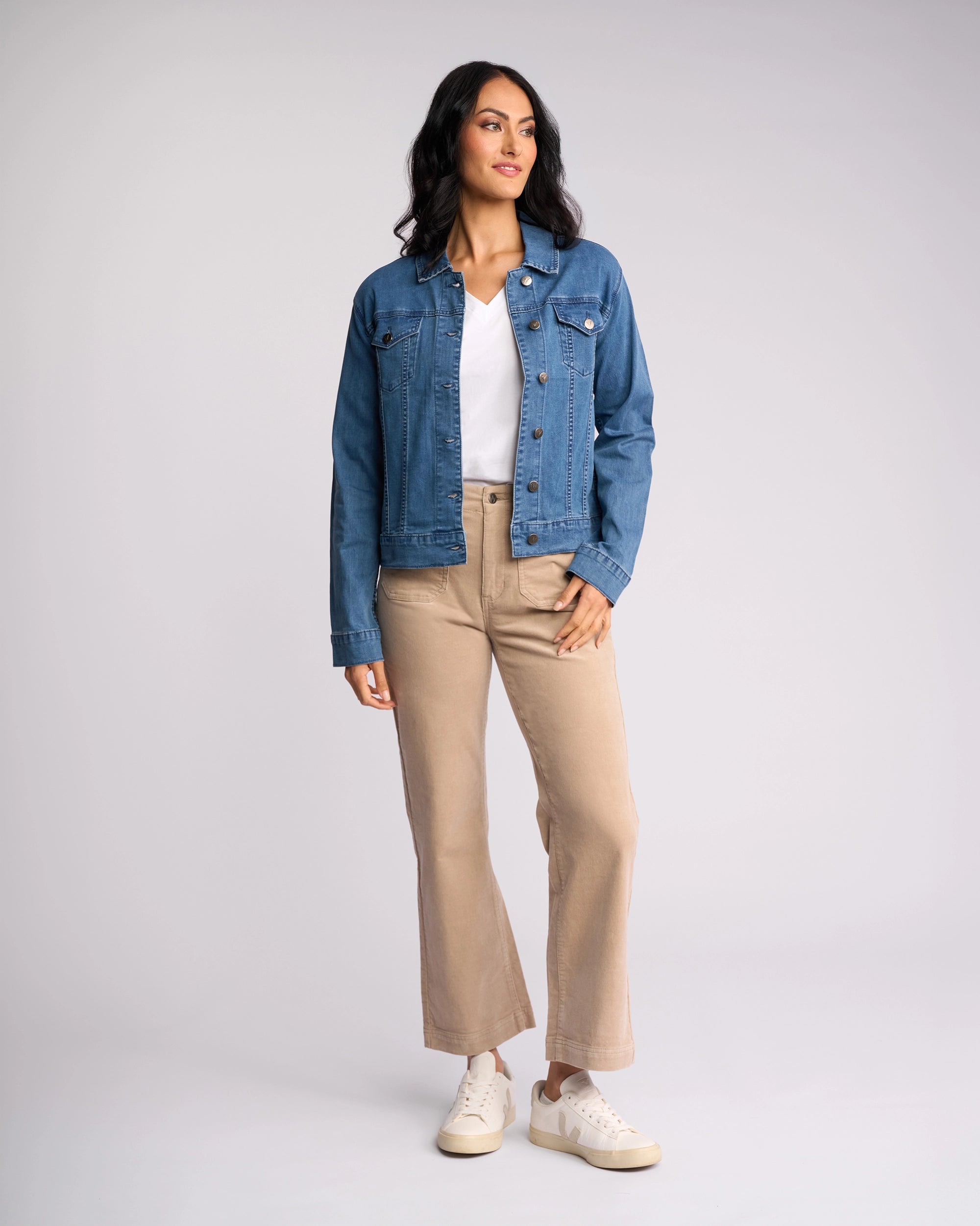 A person with long dark hair stands against a plain background wearing the Vassalli Core Jacket - Washed Denim over a white shirt, beige pants, and white sneakers, looking slightly to the side with a neutral expression.