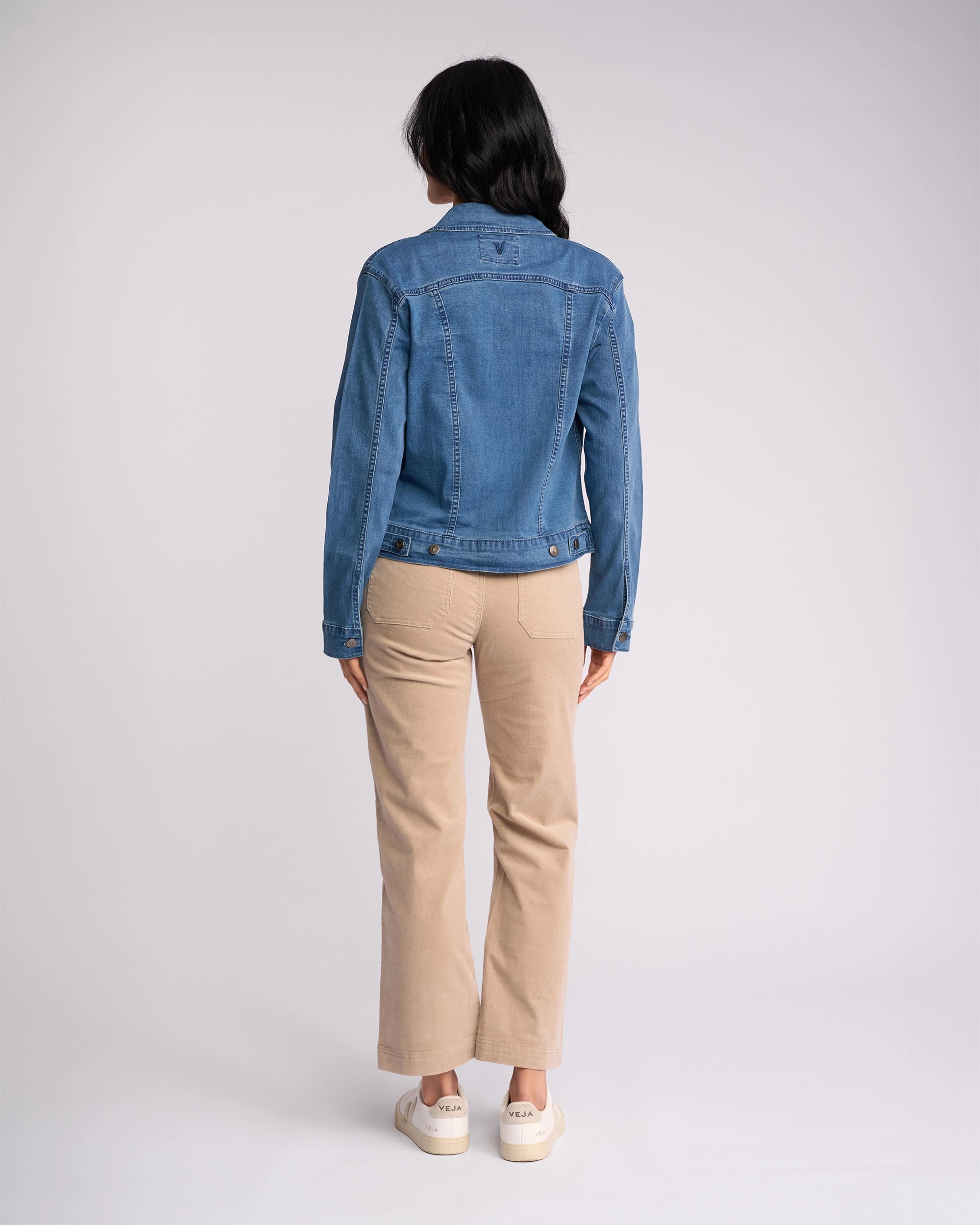 A person with long dark hair faces away, wearing the Vassalli Core Jacket - Washed Denim, beige pants, and white sneakers against a plain light background.