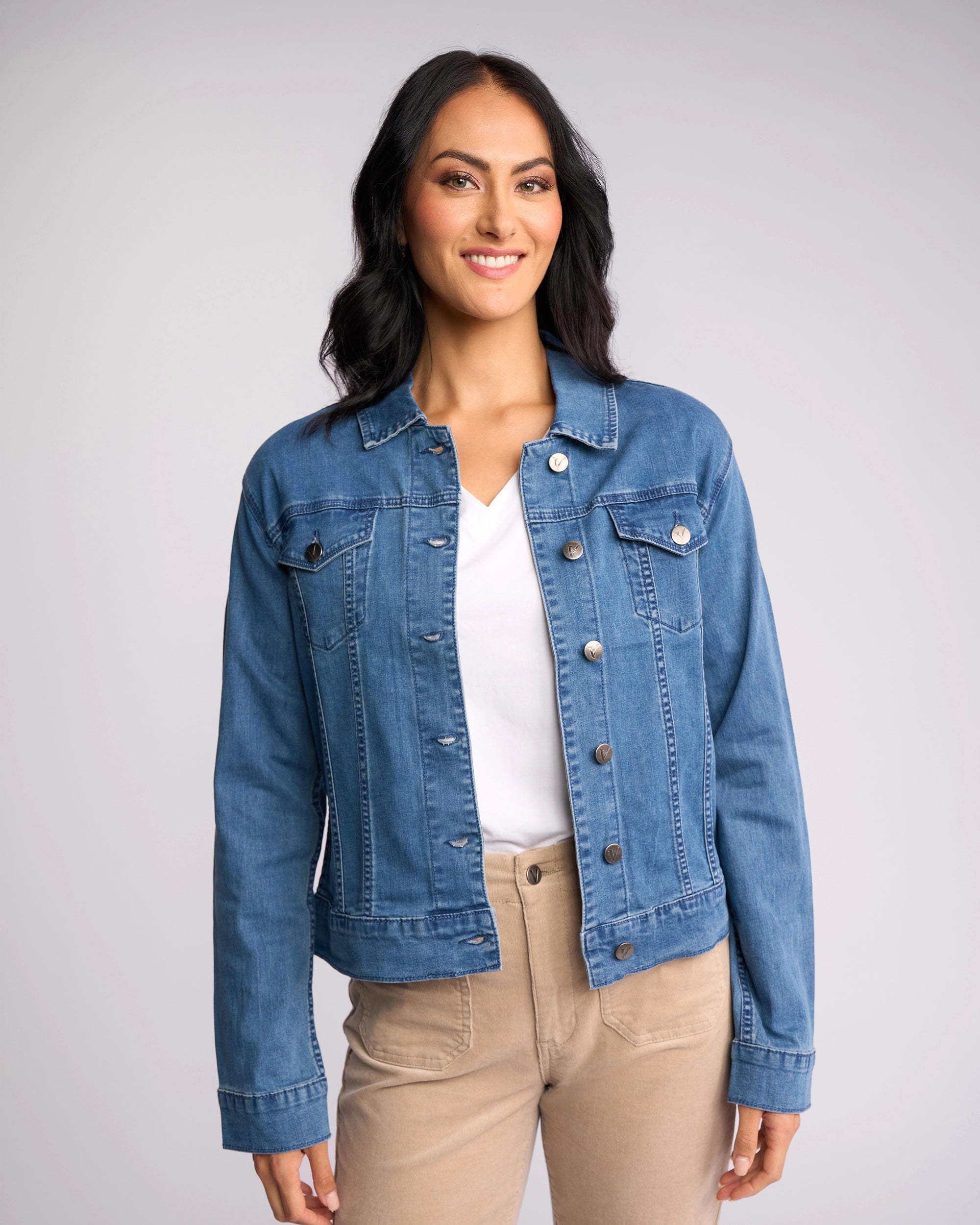 A woman with long dark hair smiles against a plain light background, wearing the Vassalli Core Jacket - Washed Denim over a white shirt and beige pants.