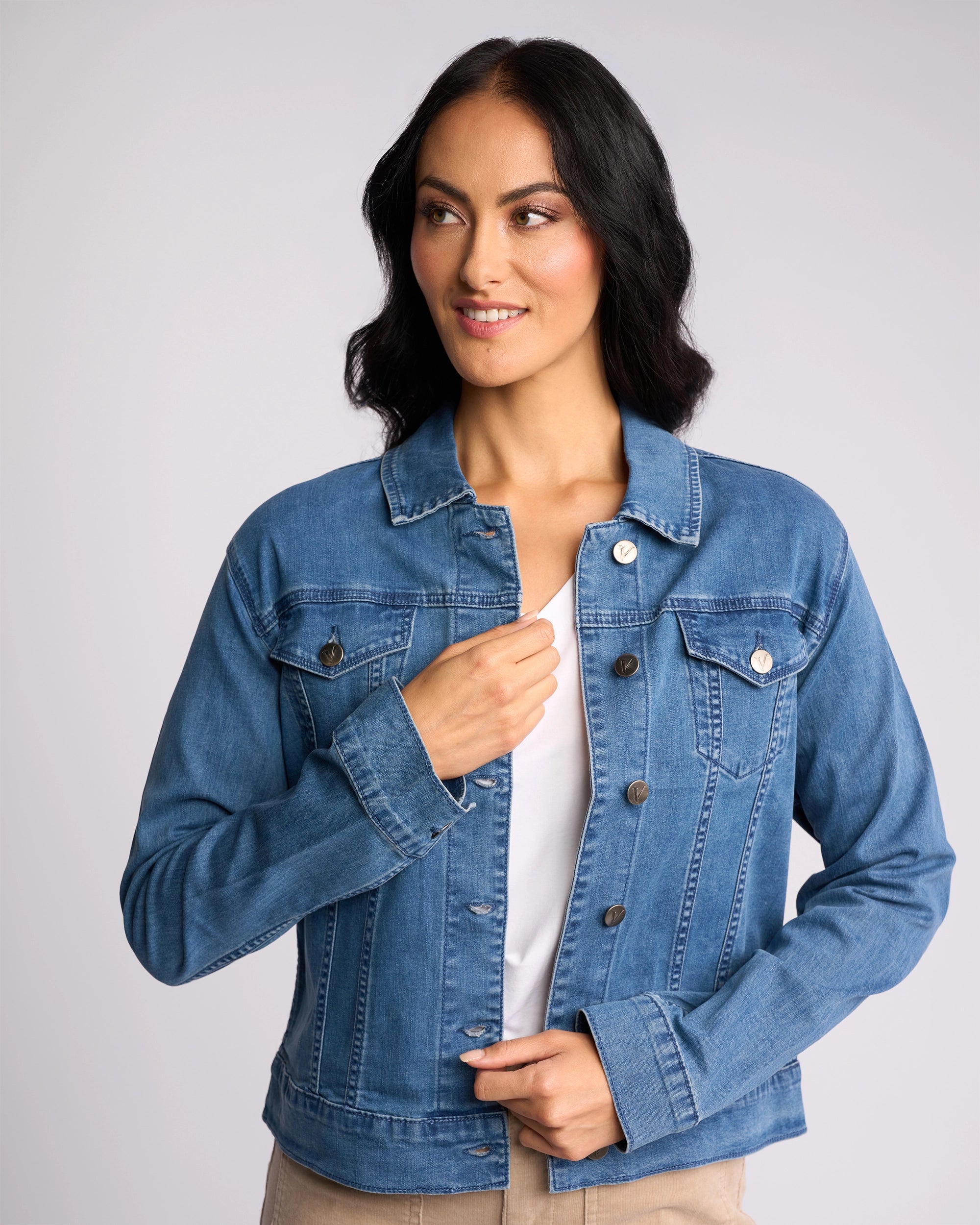 A woman with long dark hair smiles slightly and looks to the side while adjusting the front of a Vassalli Core Jacket - Washed Denim over a white shirt. The background is plain and light-colored.