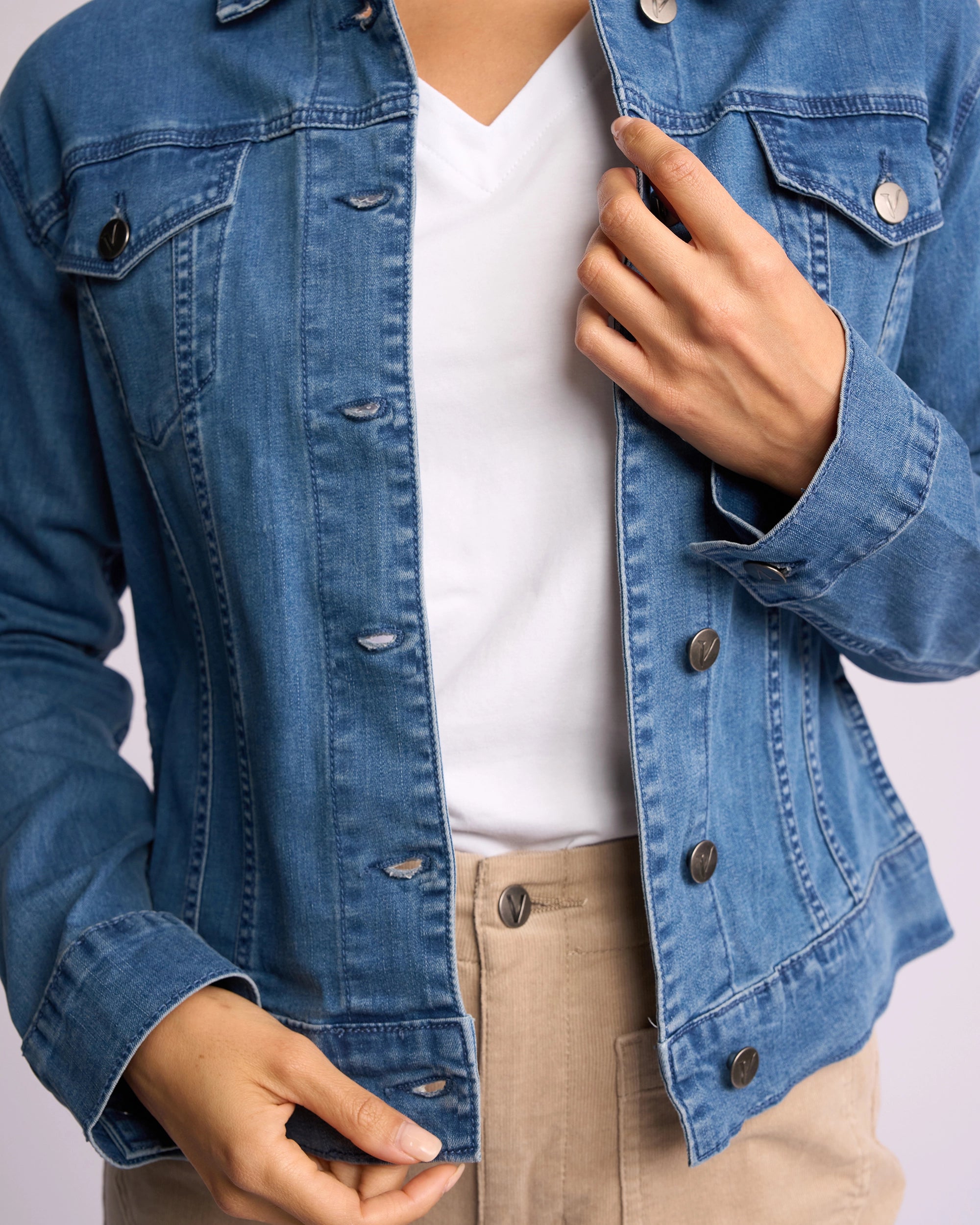 A person wearing the Vassalli Core Jacket - Washed Denim over a white shirt and beige pants, adjusting the jacket with one hand near the chest and the other at the waist. The face is not visible.