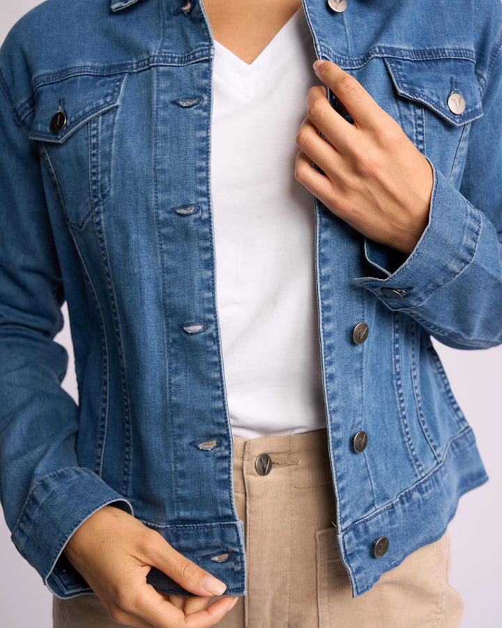 A person wearing the Vassalli Core Jacket - Washed Denim over a white shirt and beige pants, adjusting the jacket with one hand near the chest and the other at the waist. The face is not visible.