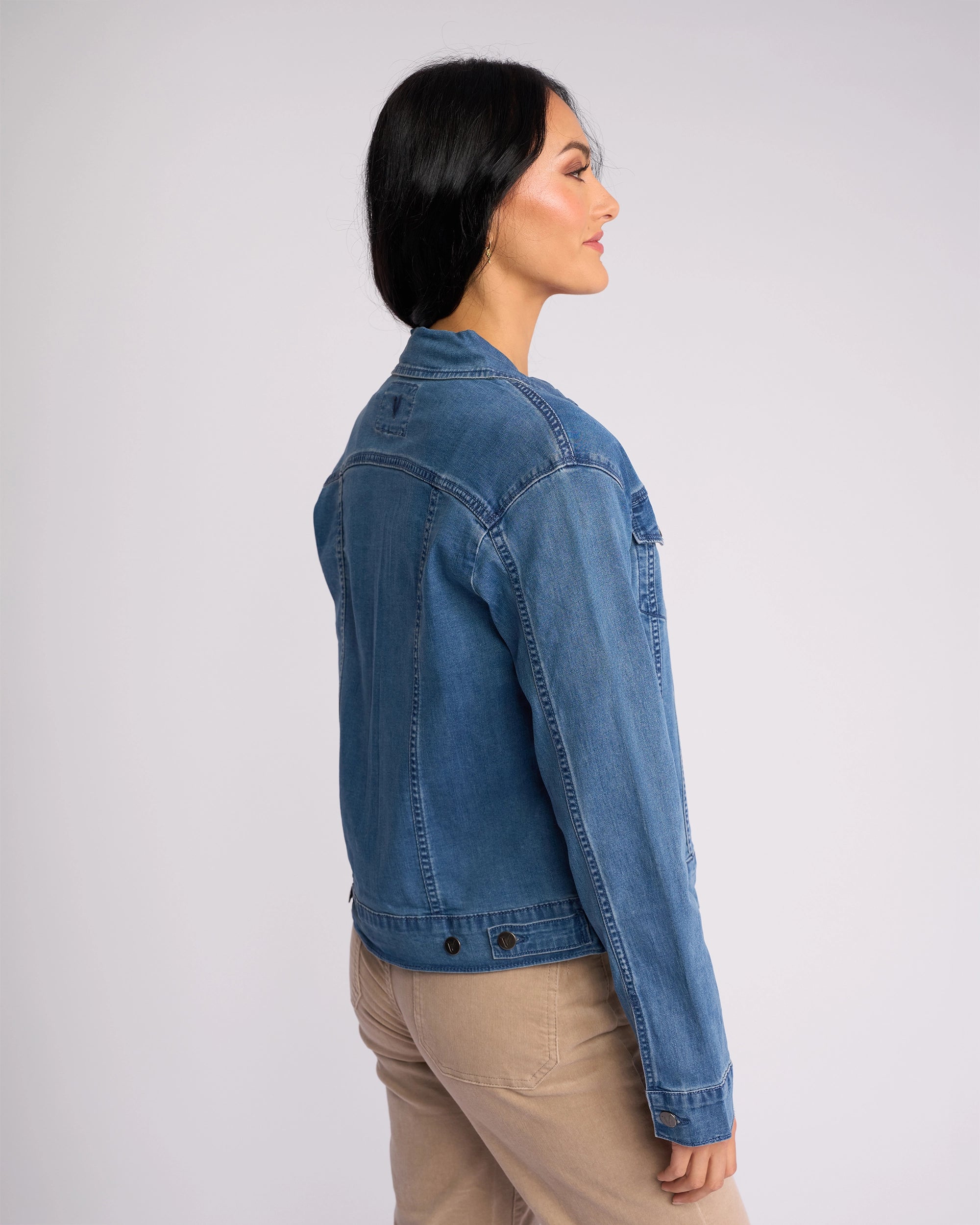 A woman with long dark hair stands sideways against a plain light background, wearing the Vassalli Core Jacket in washed denim and beige stretch denim pants.