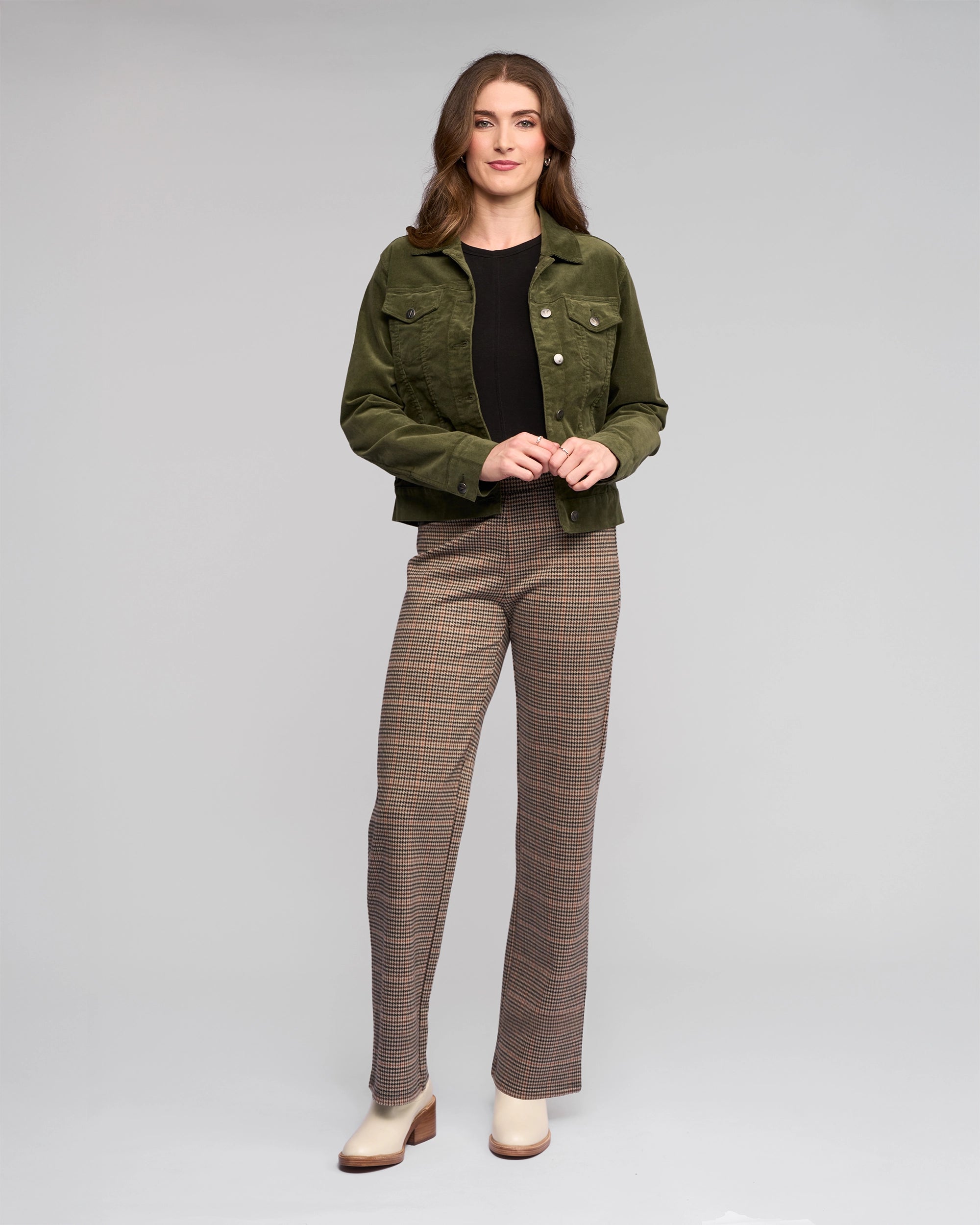 A woman stands against a plain background, wearing the Vassalli Core Cord Jacket in Deep Sage over a black top and plaid trousers with cream shoes. She smiles slightly, hands loosely clasped, embracing an effortless denim-jacket style.