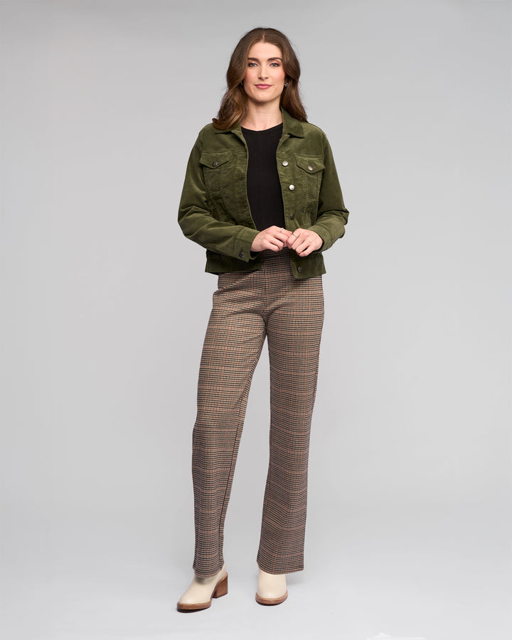 A woman stands against a plain background, wearing the Vassalli Core Cord Jacket in Deep Sage over a black top and plaid trousers with cream shoes. She smiles slightly, hands loosely clasped, embracing an effortless denim-jacket style.