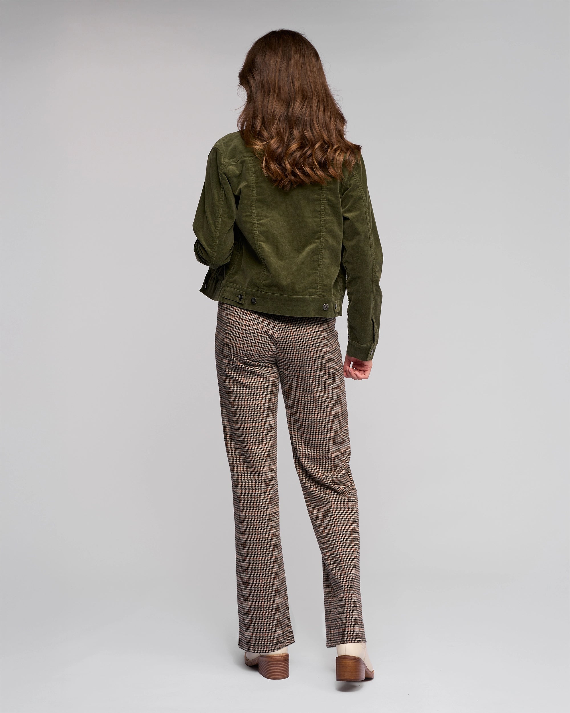 A woman with wavy brown hair stands facing away, wearing the Vassalli Core Cord Jacket in Deep Sage, styled like a denim jacket, with plaid trousers and brown-heeled shoes against a plain light gray background.