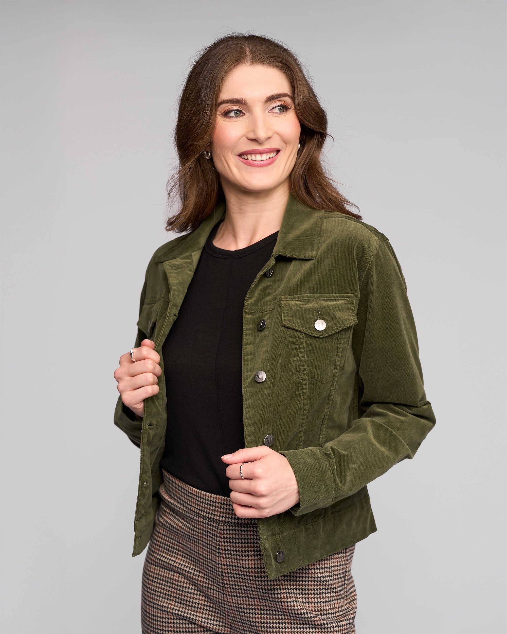 A woman with wavy brown hair smiles while looking to the side. She wears a Vassalli Core Cord Jacket in Deep Sage over a black top and checkered pants, standing against a plain light gray background.