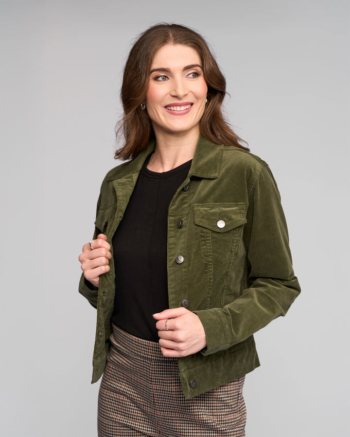 A woman with wavy brown hair smiles while looking to the side. She wears a Vassalli Core Cord Jacket in Deep Sage over a black top and checkered pants, standing against a plain light gray background.