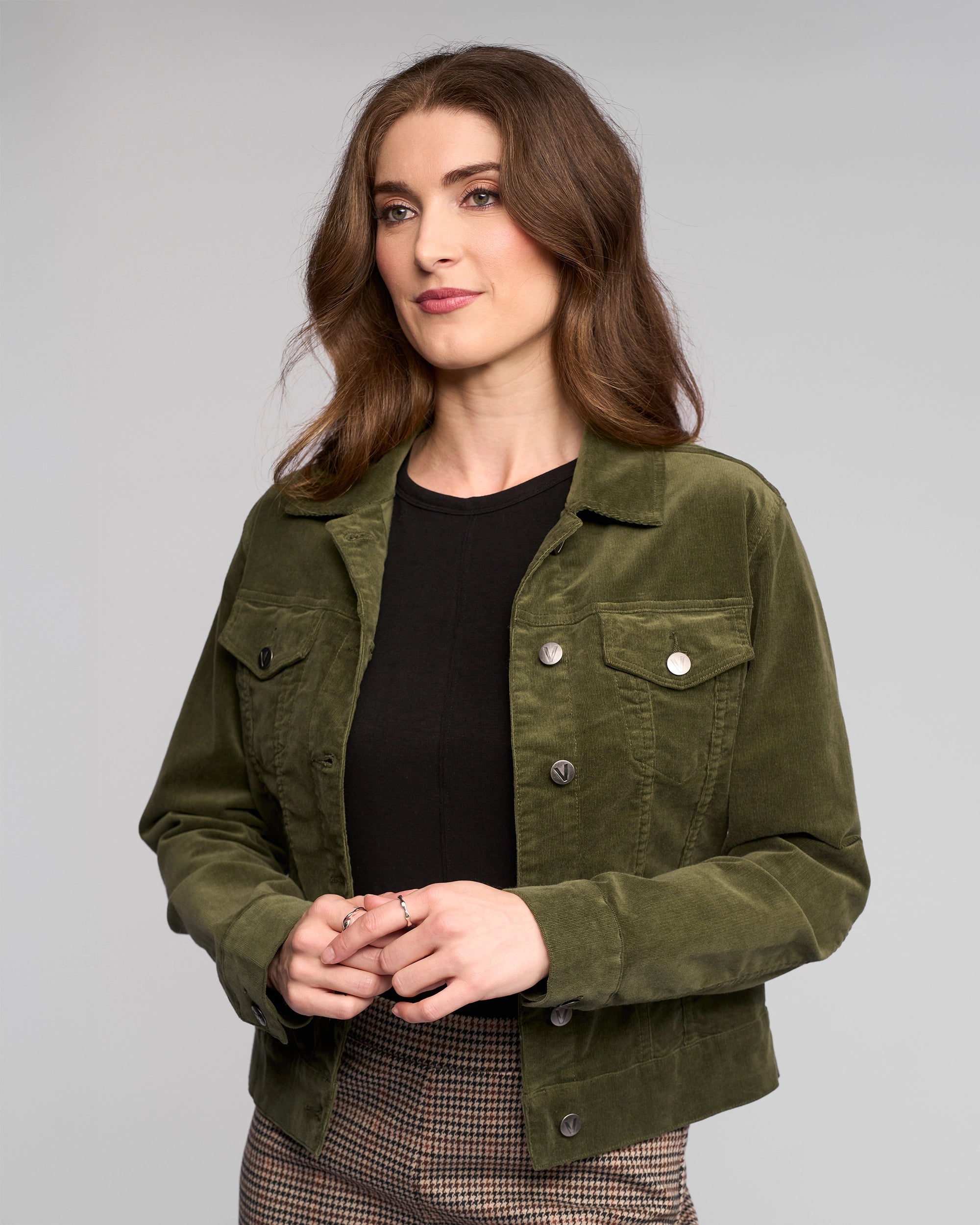 A woman with wavy brown hair wears the Vassalli Core Cord Jacket in Deep Sage over a black top and plaid pants, standing against a plain light gray background and looking slightly to the side with a soft smile.