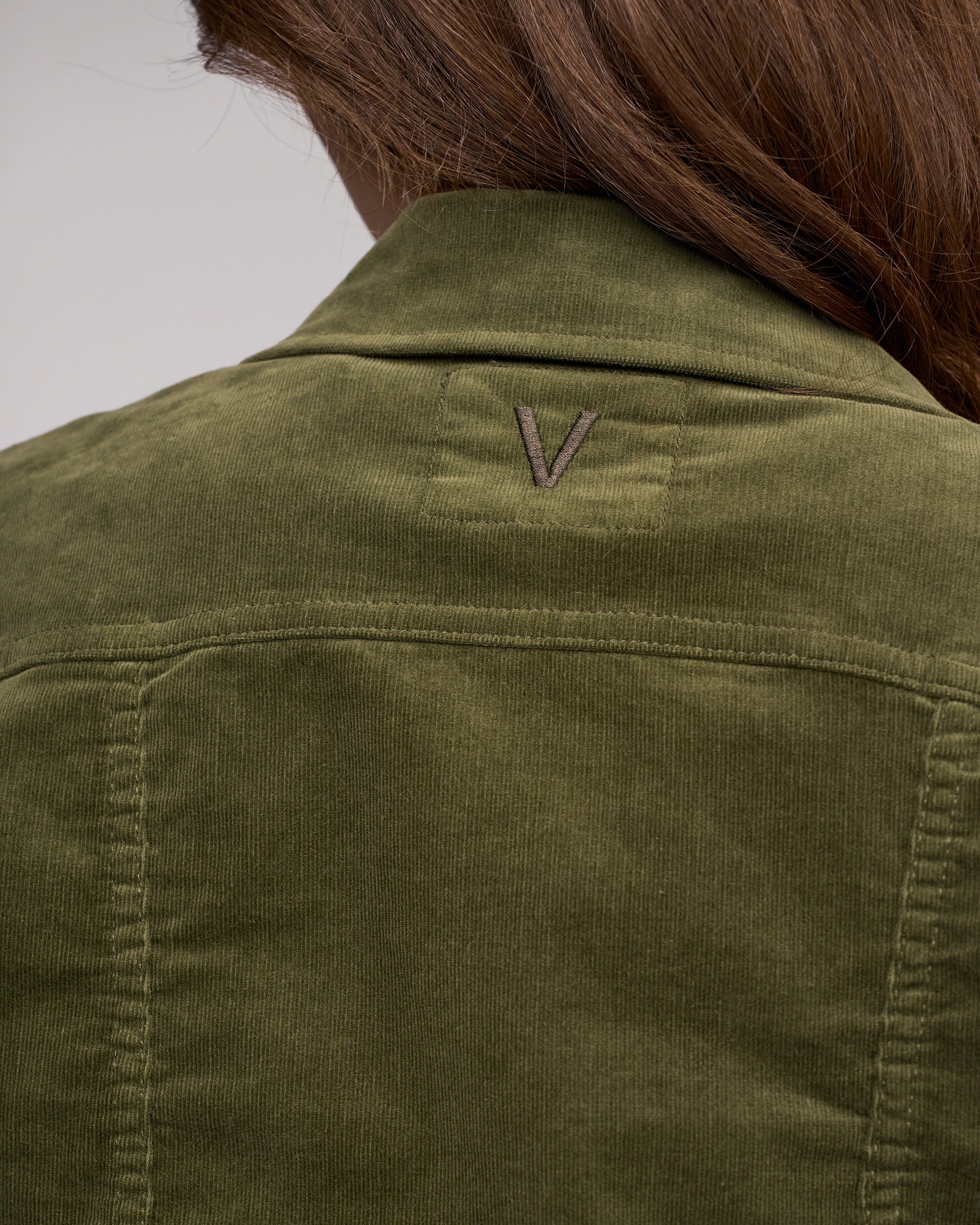 A close-up of the back of a person wearing the Vassalli Core Cord Jacket - Deep Sage, styled like a denim jacket with brown hair visible and a stitched V logo on the upper back.