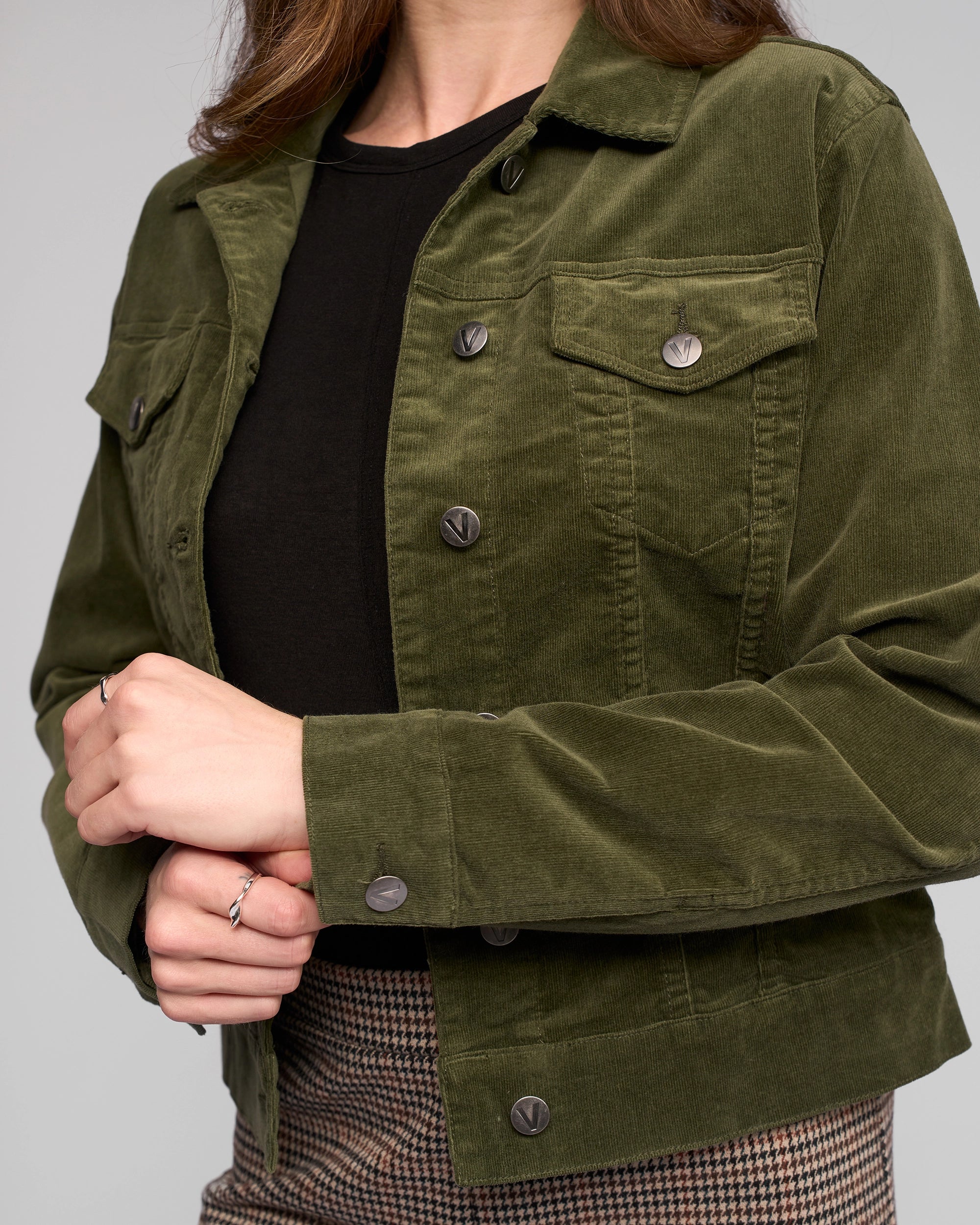 A person wears the Vassalli Core Cord Jacket - Deep Sage, styled like a denim jacket over a black top and brown plaid pants. Their hands are crossed with rings on the left hand, and the head is partially out of frame.