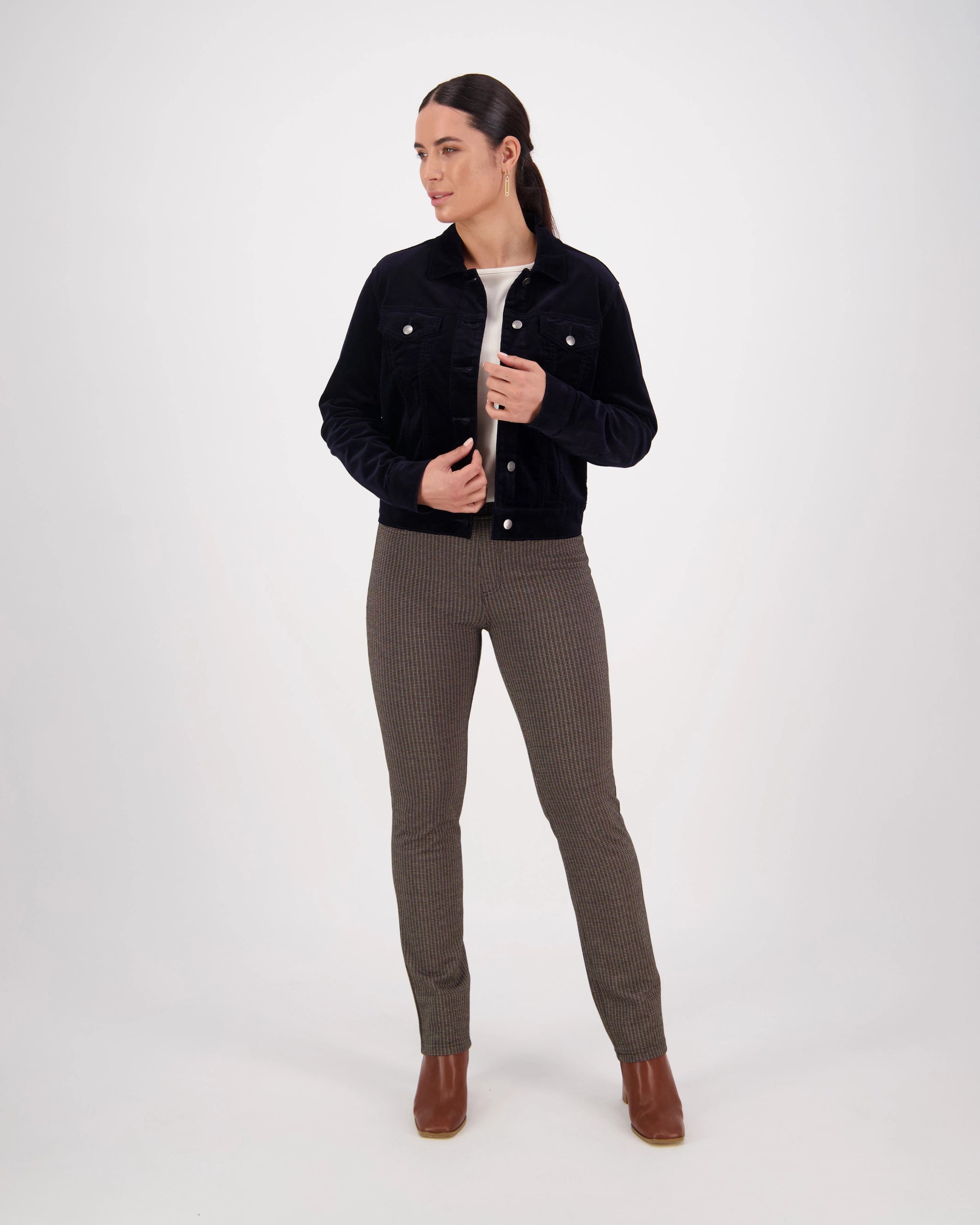 A woman stands against a plain white background wearing the Vassalli Core Cord Jacket in Midnight, styled denim-jacket fashion with a light top, checked brown pants, and brown ankle boots. Her dark hair is pulled back as she looks to the side.