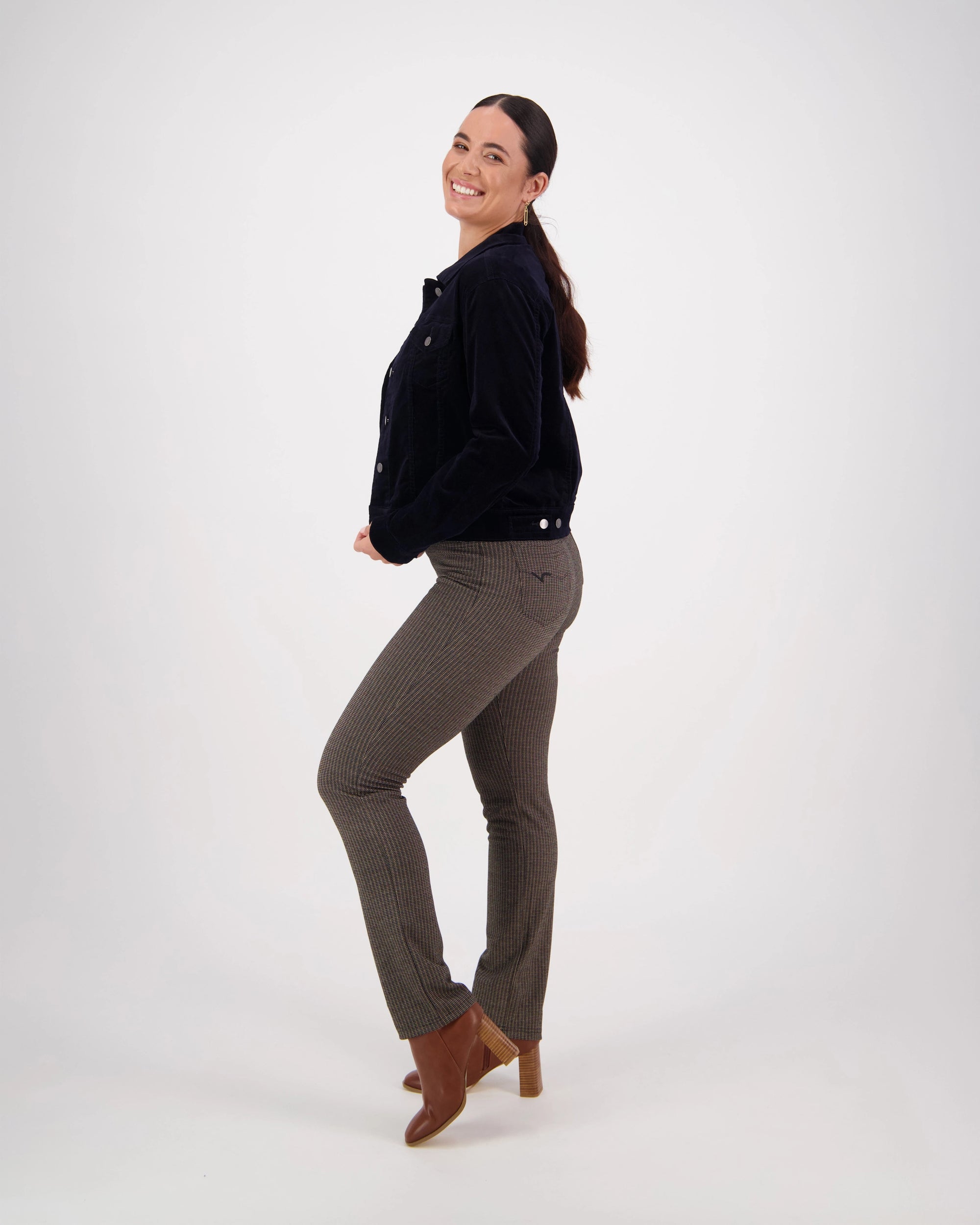 A woman with dark hair in a ponytail smiles over her shoulder, wearing the Vassalli Core Cord Jacket - Midnight, patterned pants, and brown ankle boots against a plain white background.