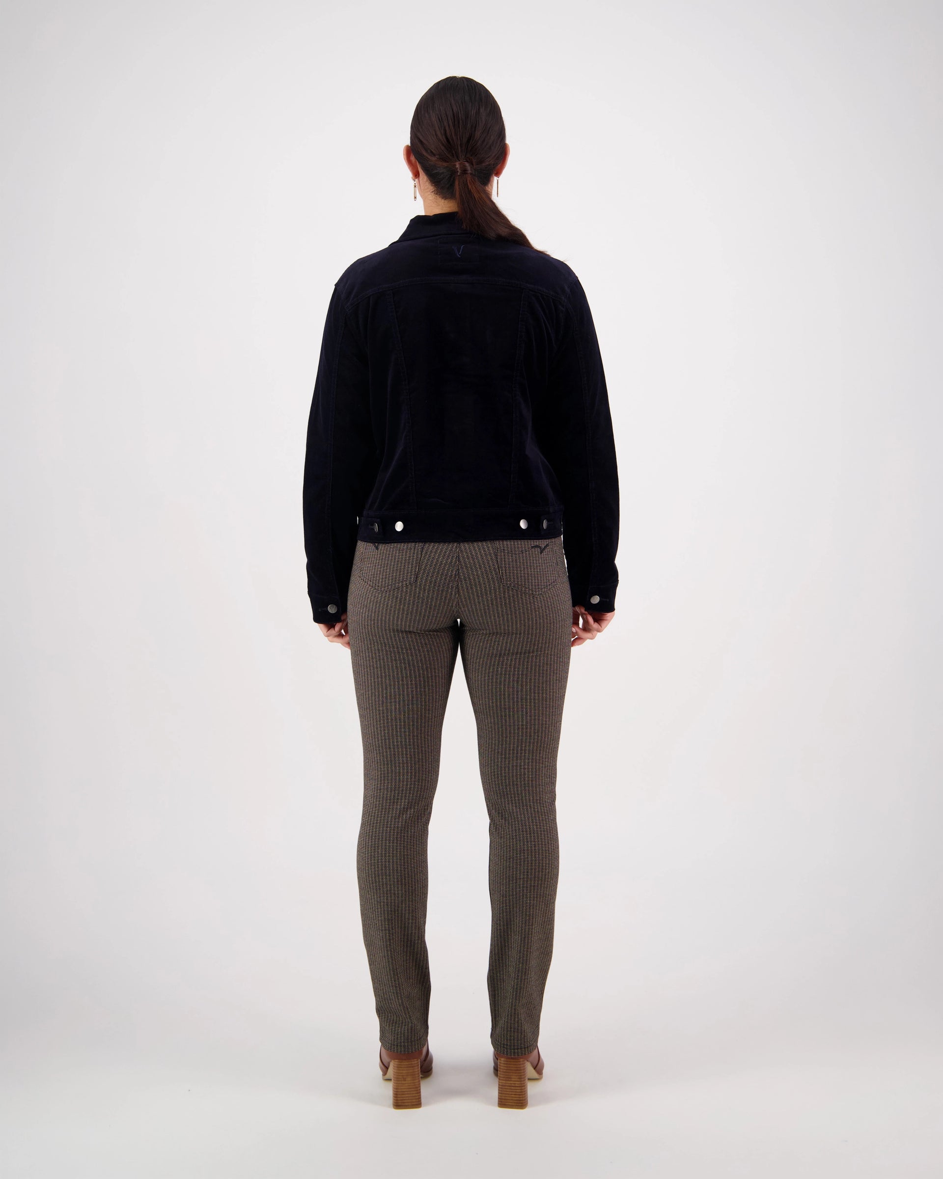 A woman stands facing away from the camera against a plain white background, wearing the Vassalli Core Cord Jacket - Midnight—a timeless wardrobe staple.