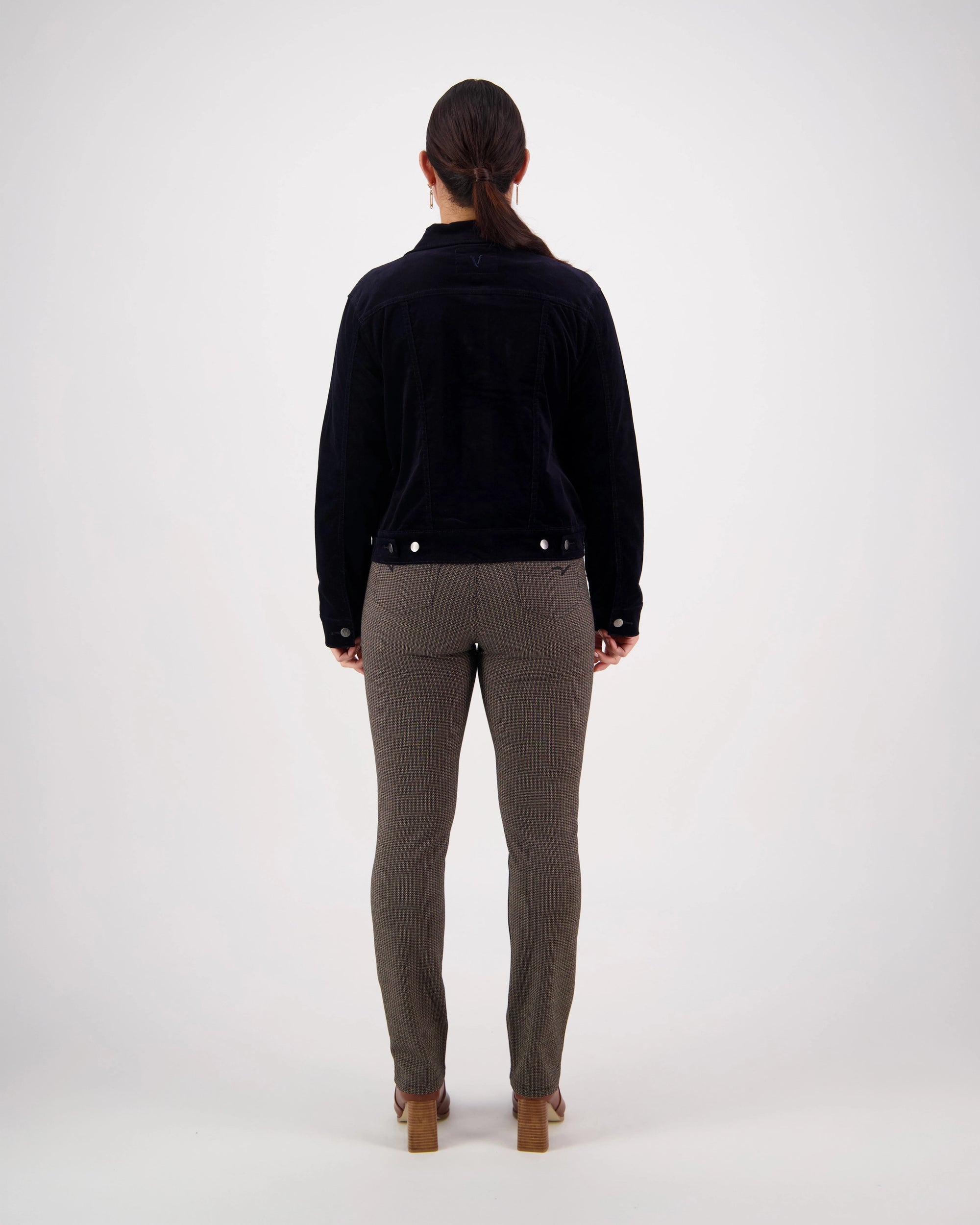 A woman stands facing away from the camera against a plain white background, wearing the Vassalli Core Cord Jacket - Midnight—a timeless wardrobe staple.