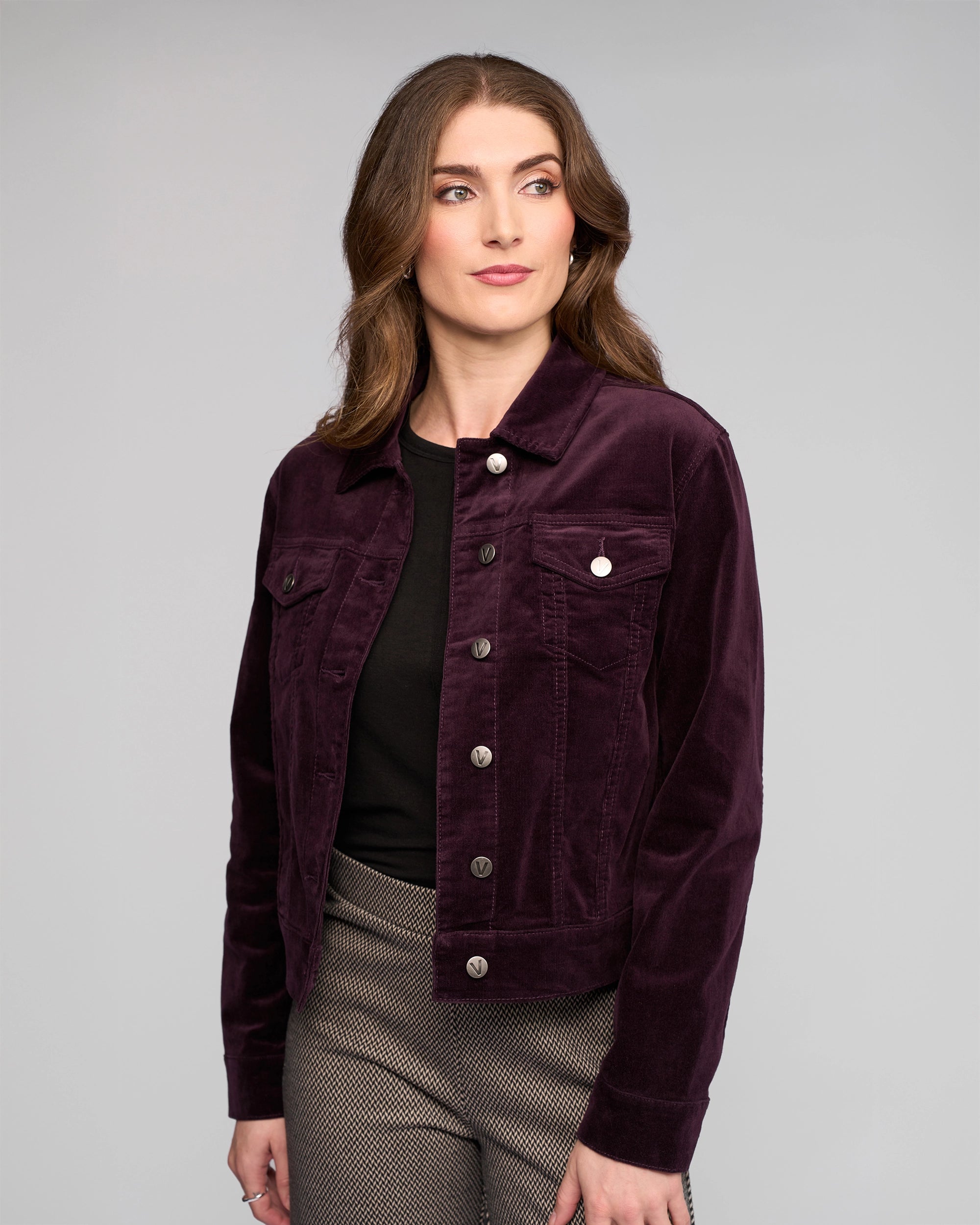 A woman with wavy brown hair wears the Vassalli Core Cord Jacket in Mulberry over a black top and patterned gray pants, looking slightly to the side against a plain gray background.
