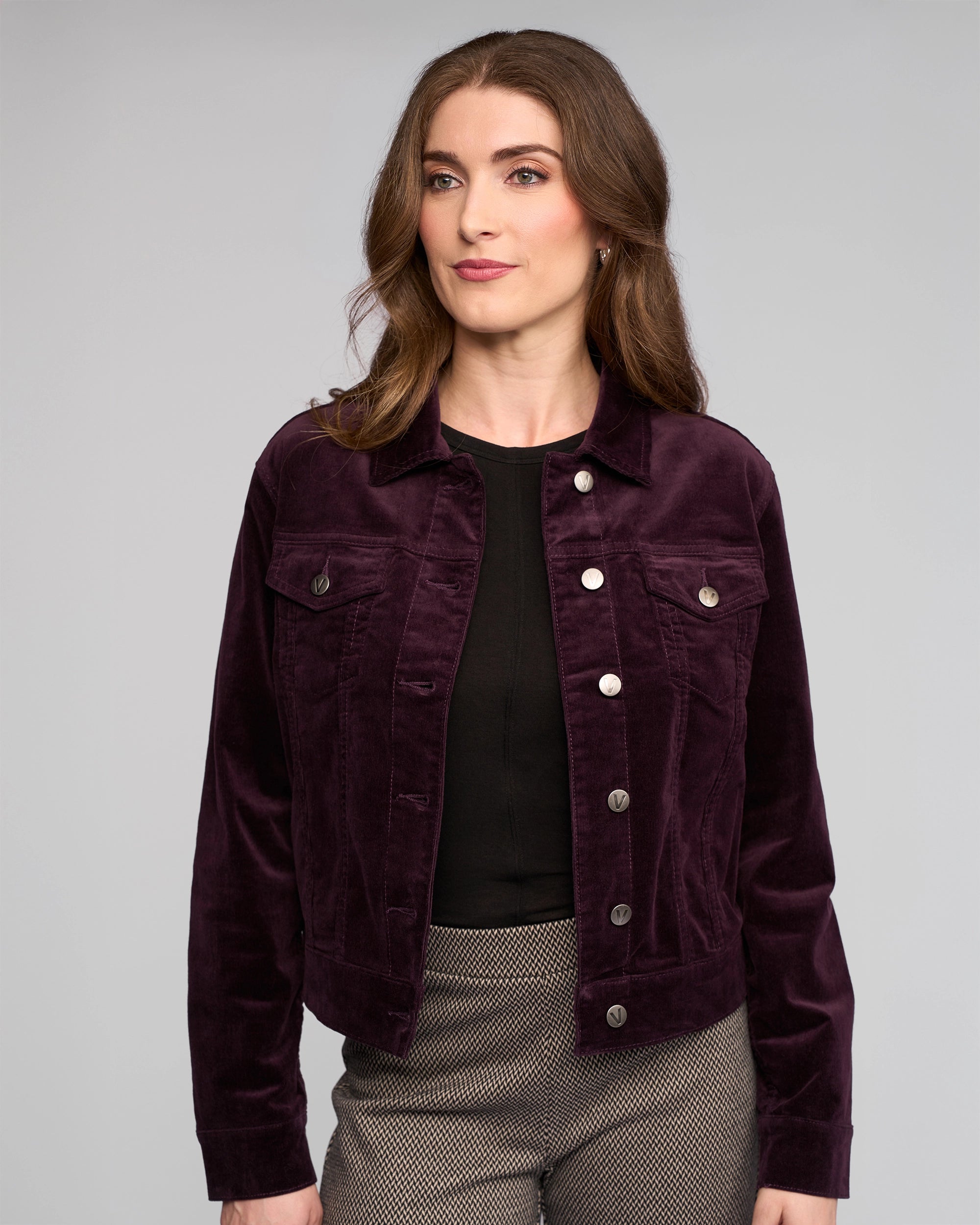 A woman with wavy brown hair models the Vassalli Core Cord Jacket in Mulberry, styled denim-jacket fashion over a black top and patterned pants, standing against a plain light gray background.
