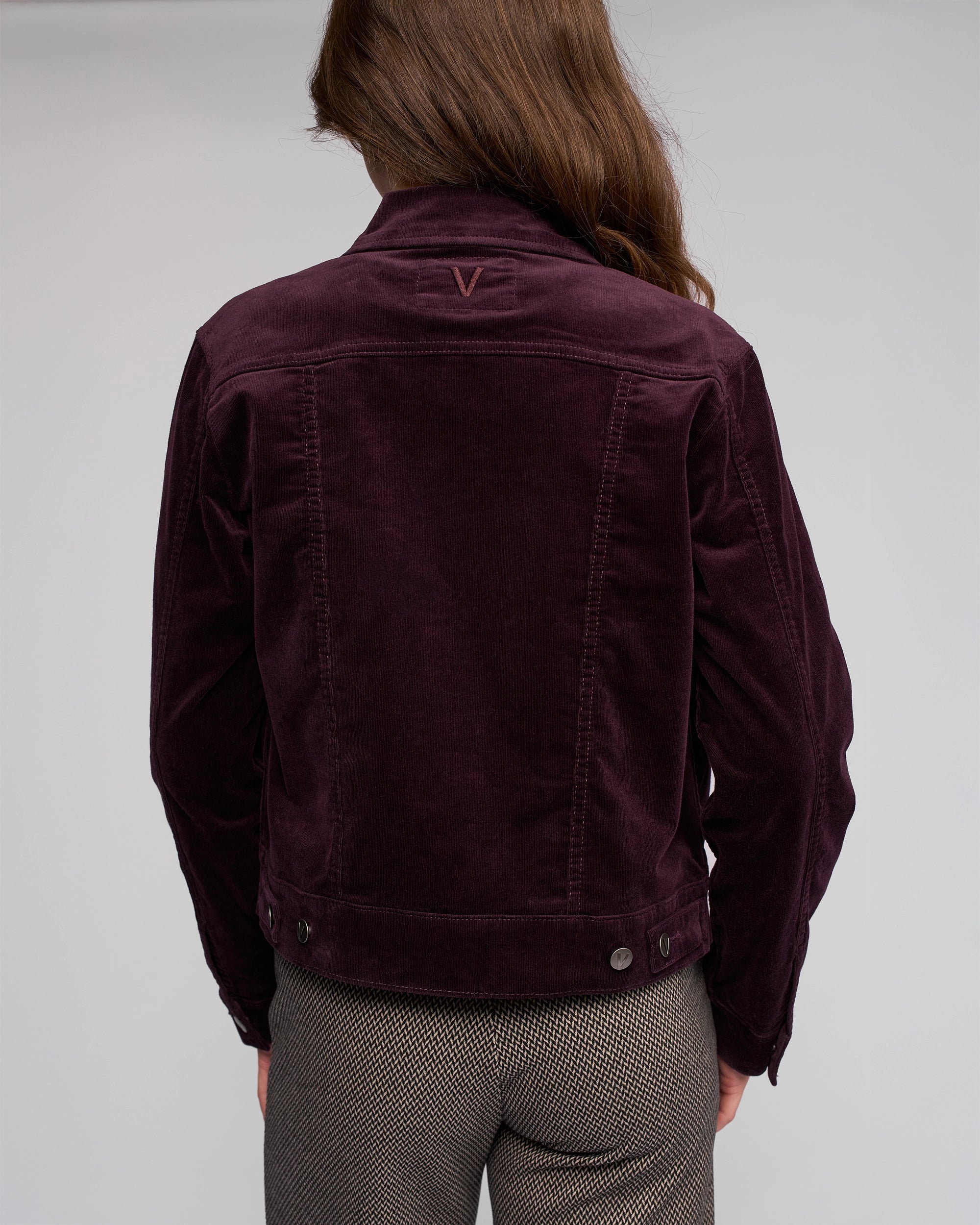 A person with long brown hair is seen from behind wearing the Vassalli Core Cord Jacket in mulberry, featuring a denim-jacket style with V stitching, paired with gray patterned pants against a plain light gray background.